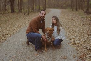 A happy dog in a plaid bandana plays with its owners on a trail covered in fall leaves.