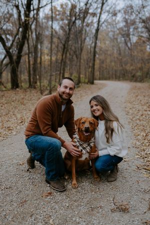 A couple and their dog share a playful moment on a leaf-covered path in the woods.