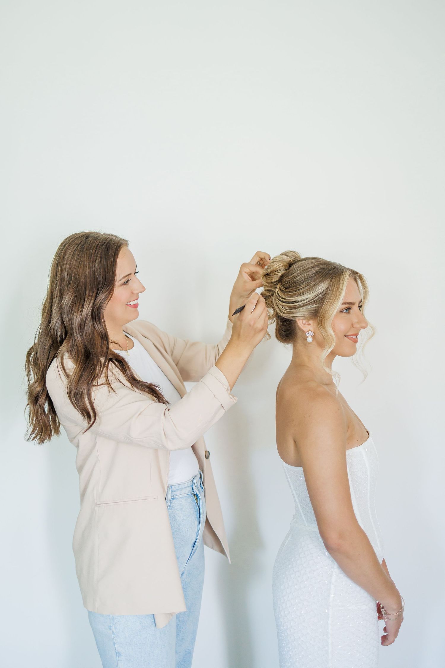 A hairstylist in a beige blazer and jeans styles an elegant updo on a woman wearing a white strapless dress.