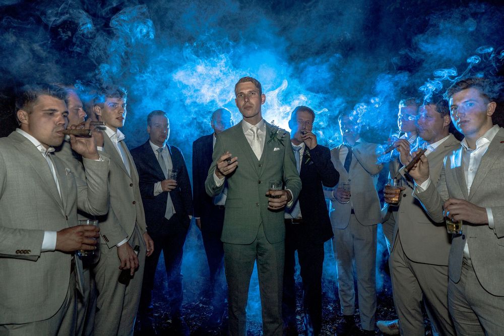 Groom and groomsmen in suits, smoking cigars and holding drinks, surrounded by dramatic blue smoke.