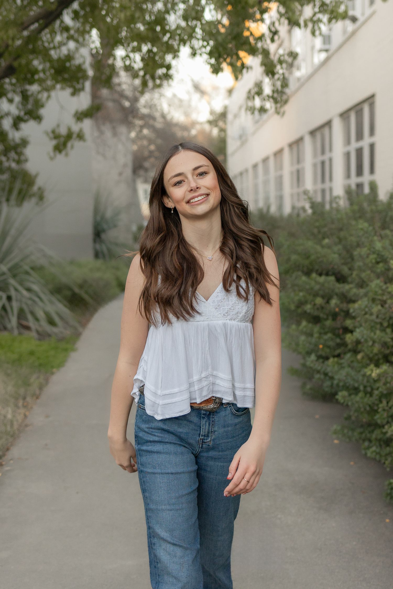 UC Davis grad photo of white female student with long brown hair, white tank, and jeans walking toward the camera with a smile