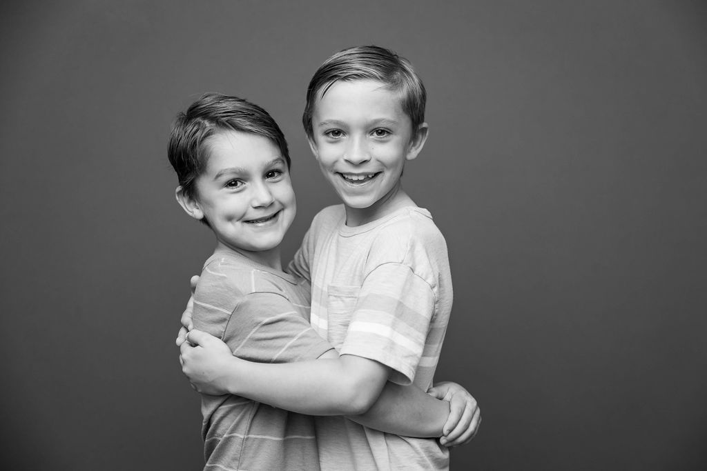 A black and white studio portrait of two young children hugging and smiling against a dark background.