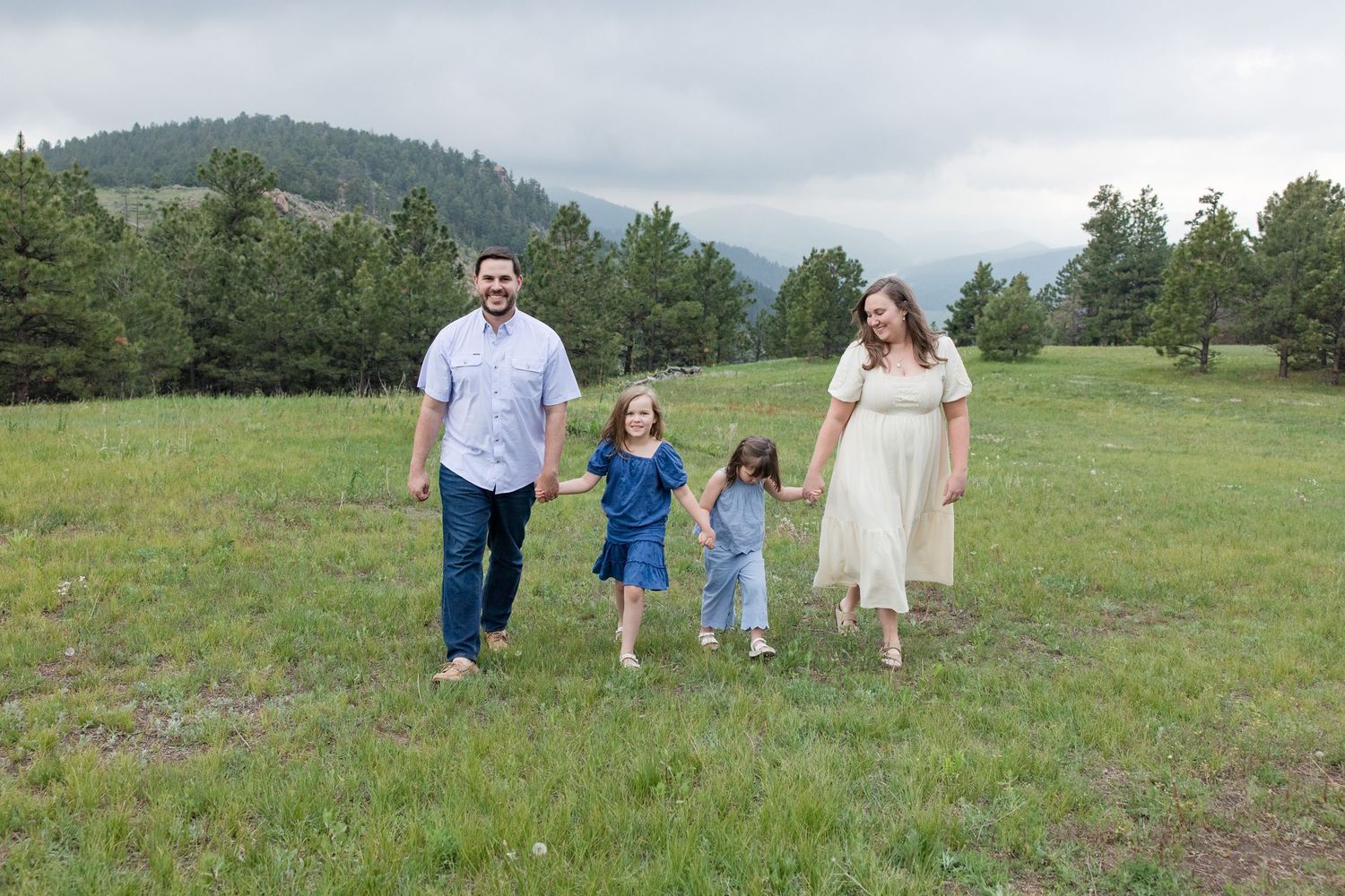 Four people walking together through a grassy meadow with pine trees and mountains in the background.