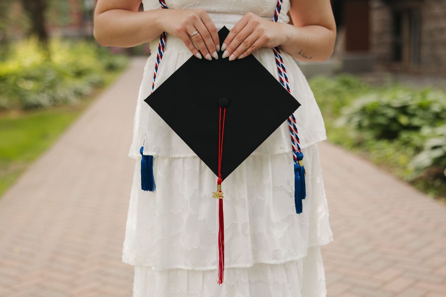 A graduate wearing a white dress holds a black graduation cap with red tassel and blue tassels on the corners.