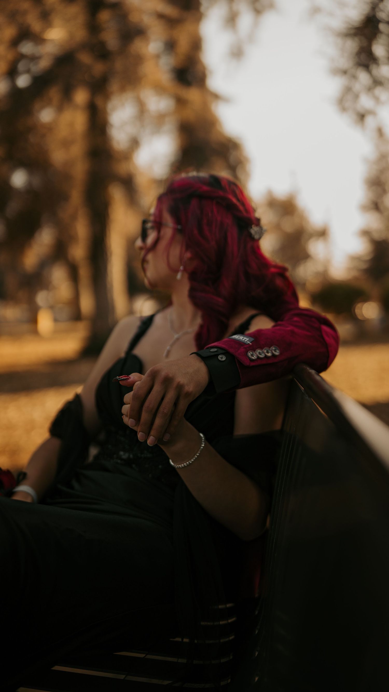 Person with vibrant red hair sitting on a park bench at sunset wearing a black outfit and Crocs shoes.