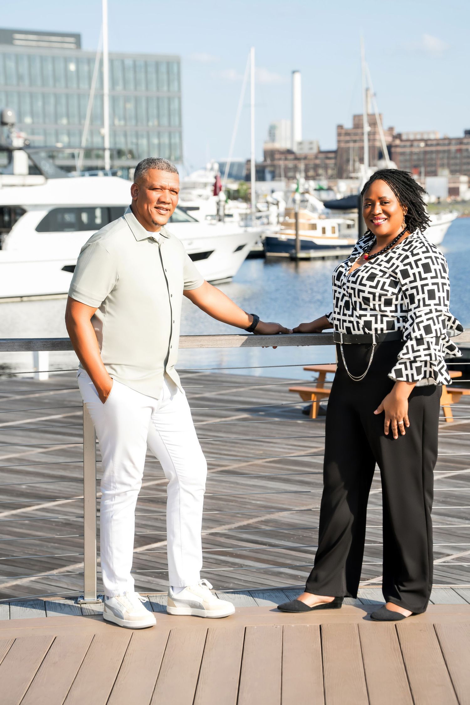 Two people stand together on a wooden dock at a marina with boats and buildings in the background.