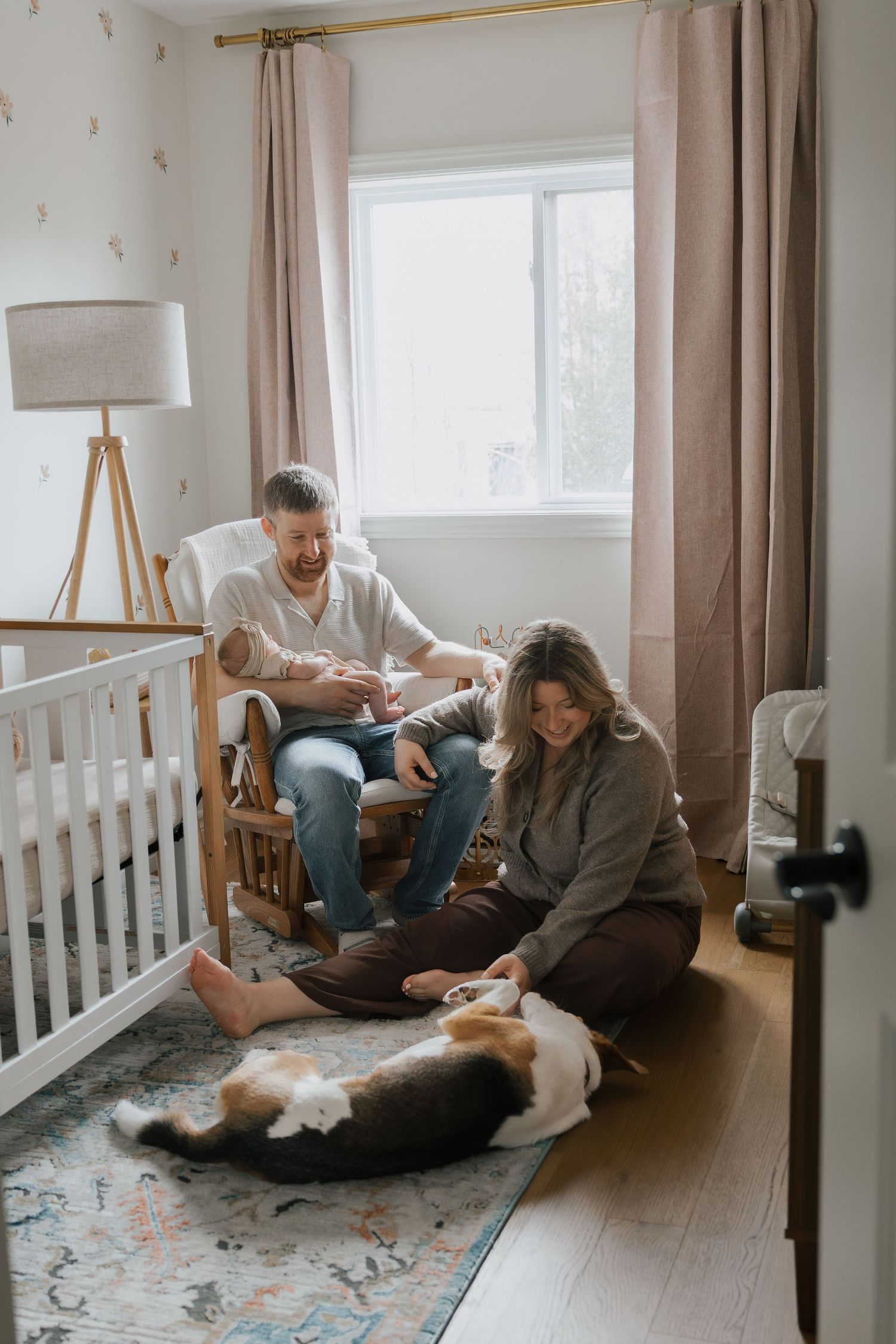 New parents with newborn baby in nursery, father in rocking chair while mother sits on floor with family dog.