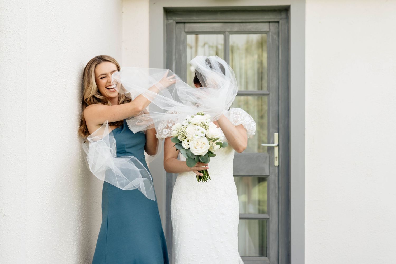 Two people share a joyful moment with white flowers by a grey door.