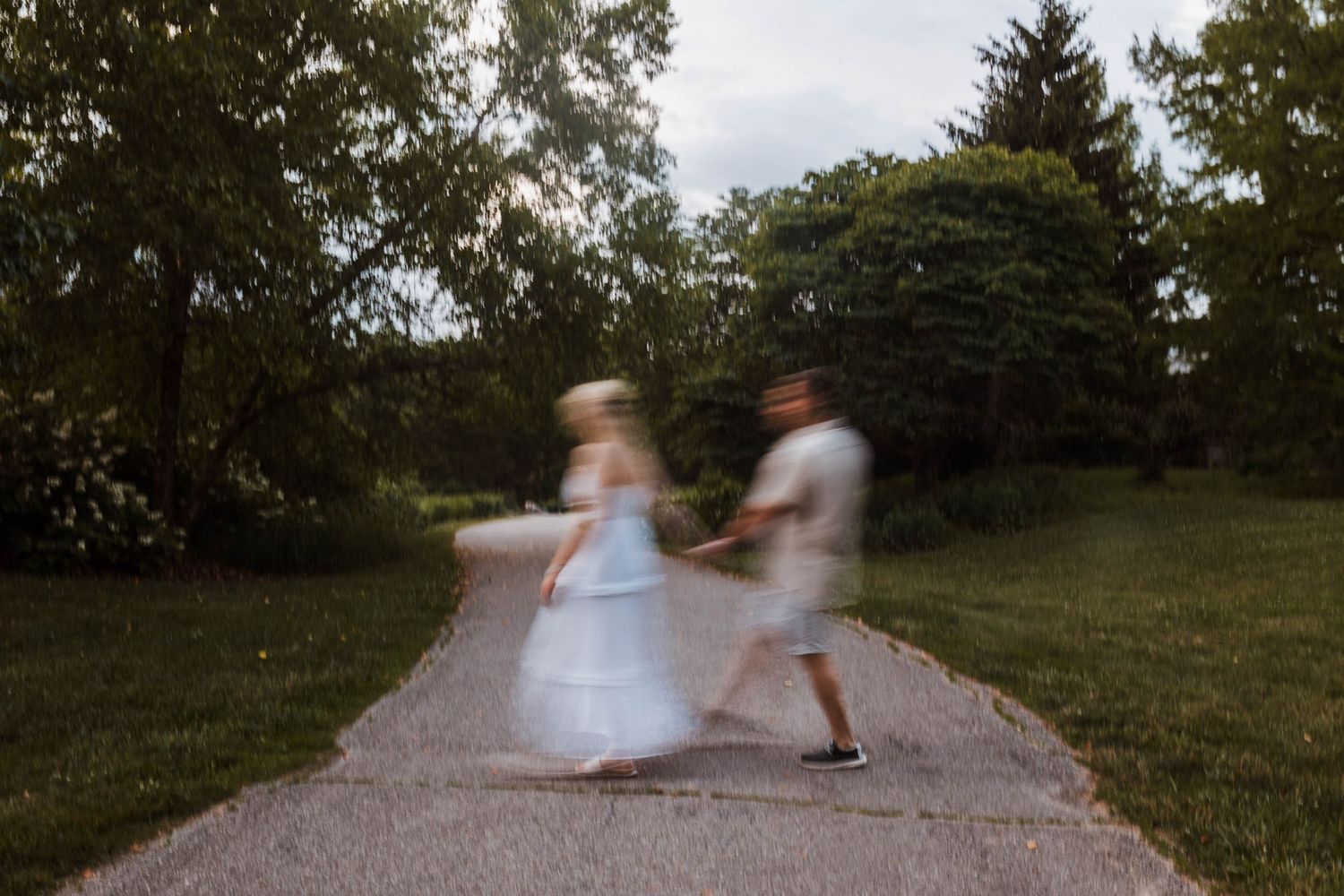 Motion blur photo of couple spinning and dancing together on a garden path surrounded by trees.