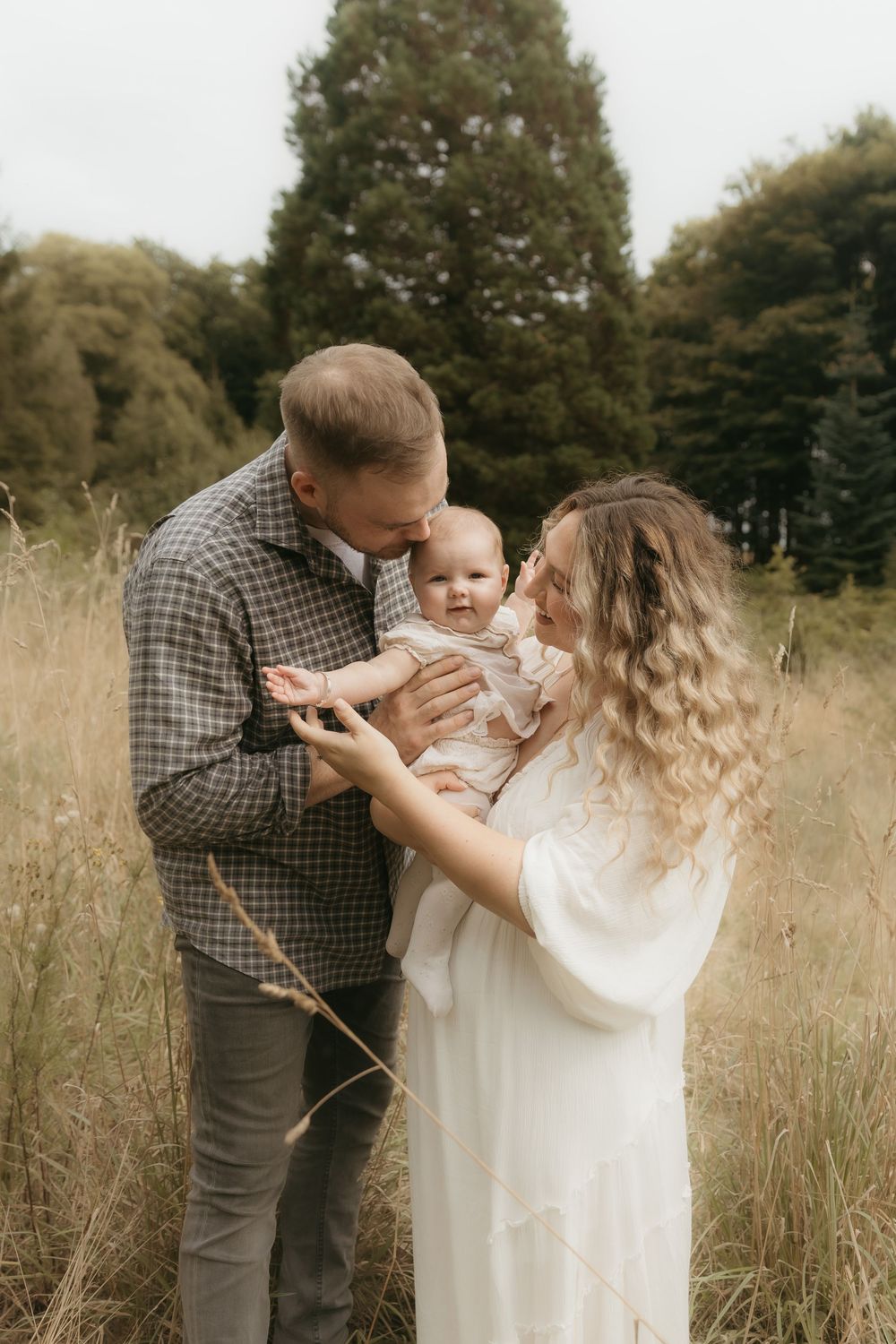 A family stands together in a rustic field with evergreen trees behind them during a portrait session.
