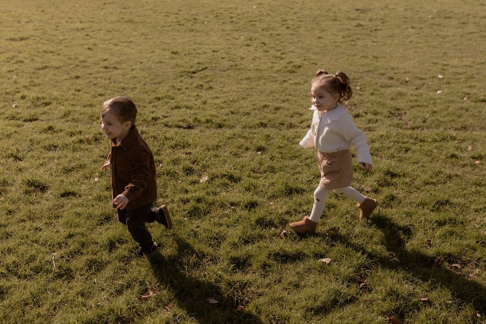 Two young children run playfully across a sunlit grassy field casting long shadows in the golden hour light.