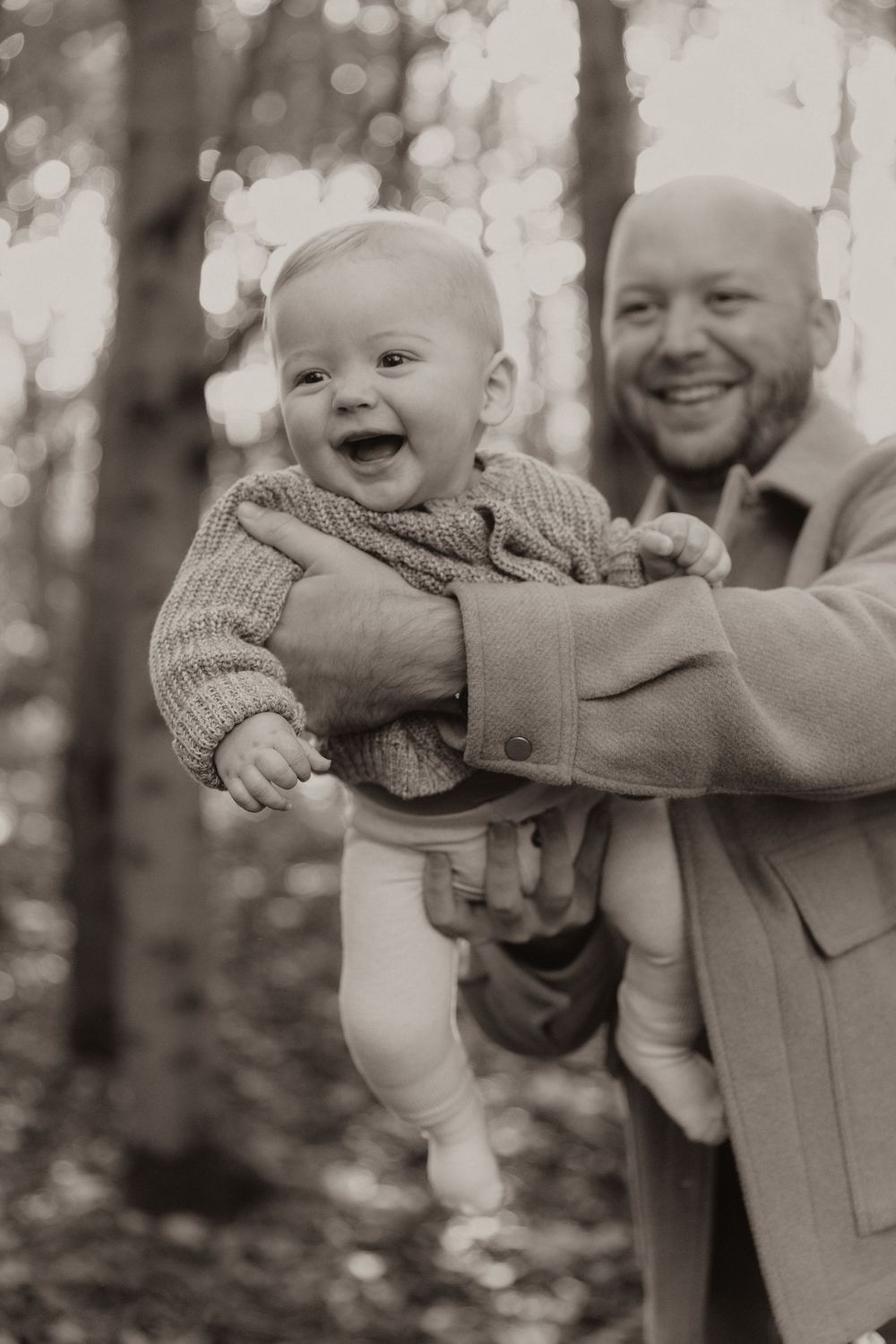 Black and white portrait of a joyful baby in a sweater being held up outdoors with trees in the background.