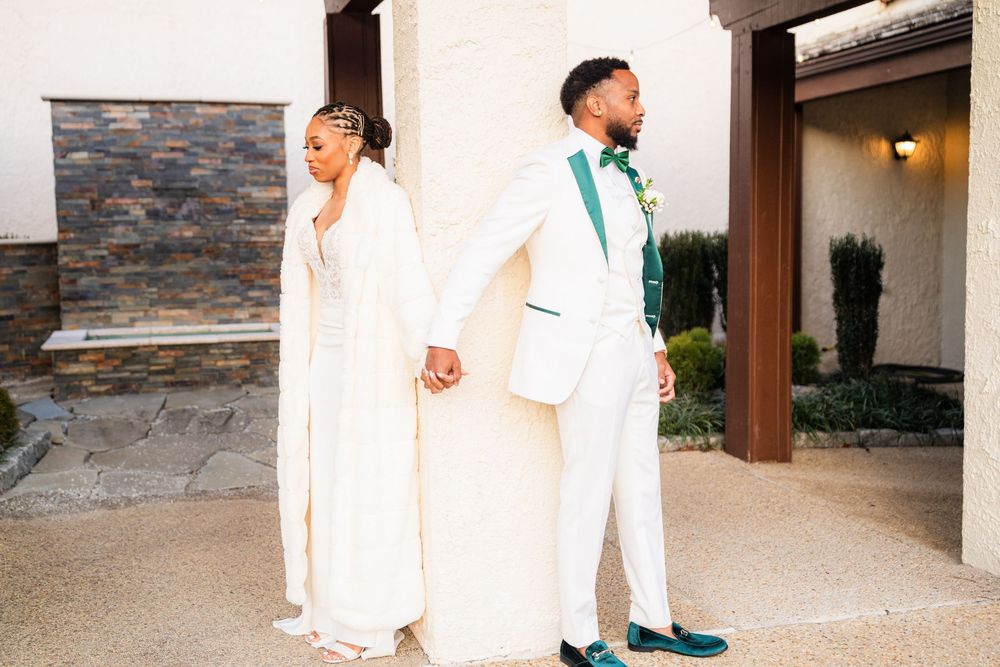 A couple in elegant white wedding attire stands together holding hands on a paved walkway outside a stone building.