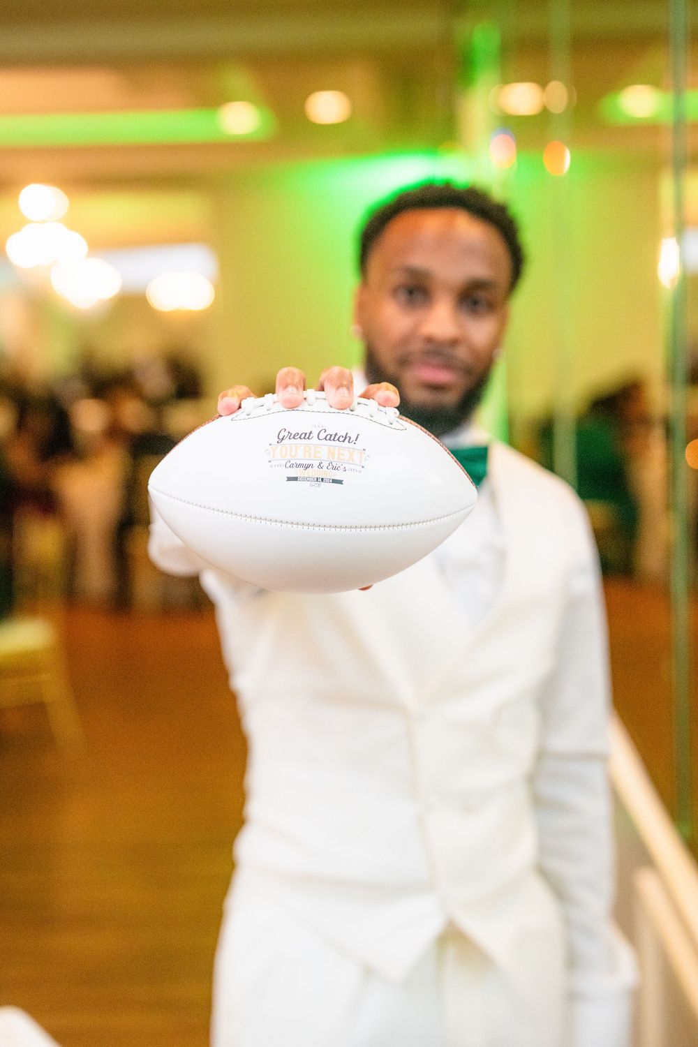 Someone in a white suit holds up a white football in an elegantly lit indoor venue.