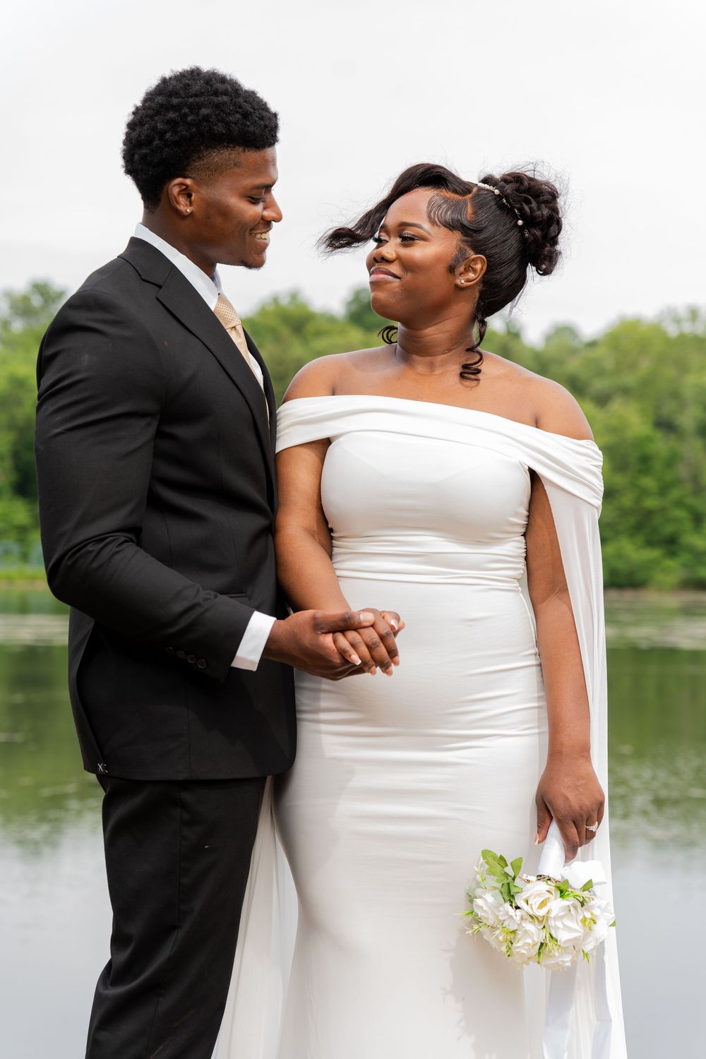 Elegant couple in wedding attire smile lovingly at each other by a serene lake during their outdoor wedding photoshoot.
