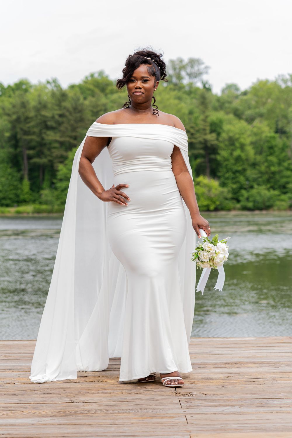 An elegant bride poses in an off-shoulder white wedding gown with cape sleeves on a wooden dock by a serene lake.