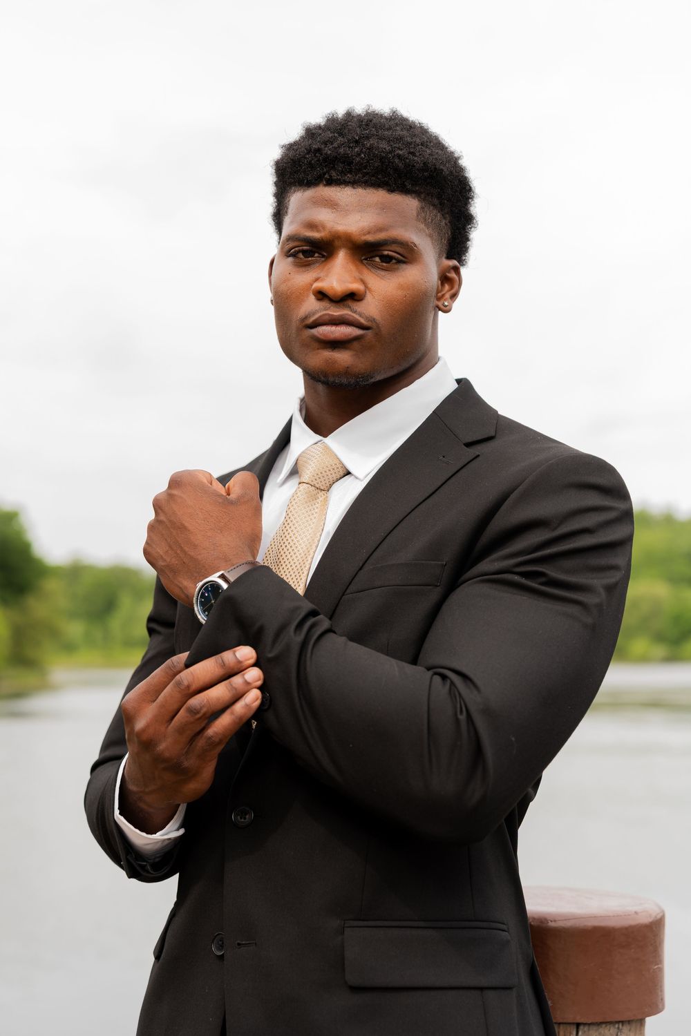 Professional portrait shot of someone in a black suit and gold tie adjusting their cuff by a waterfront.