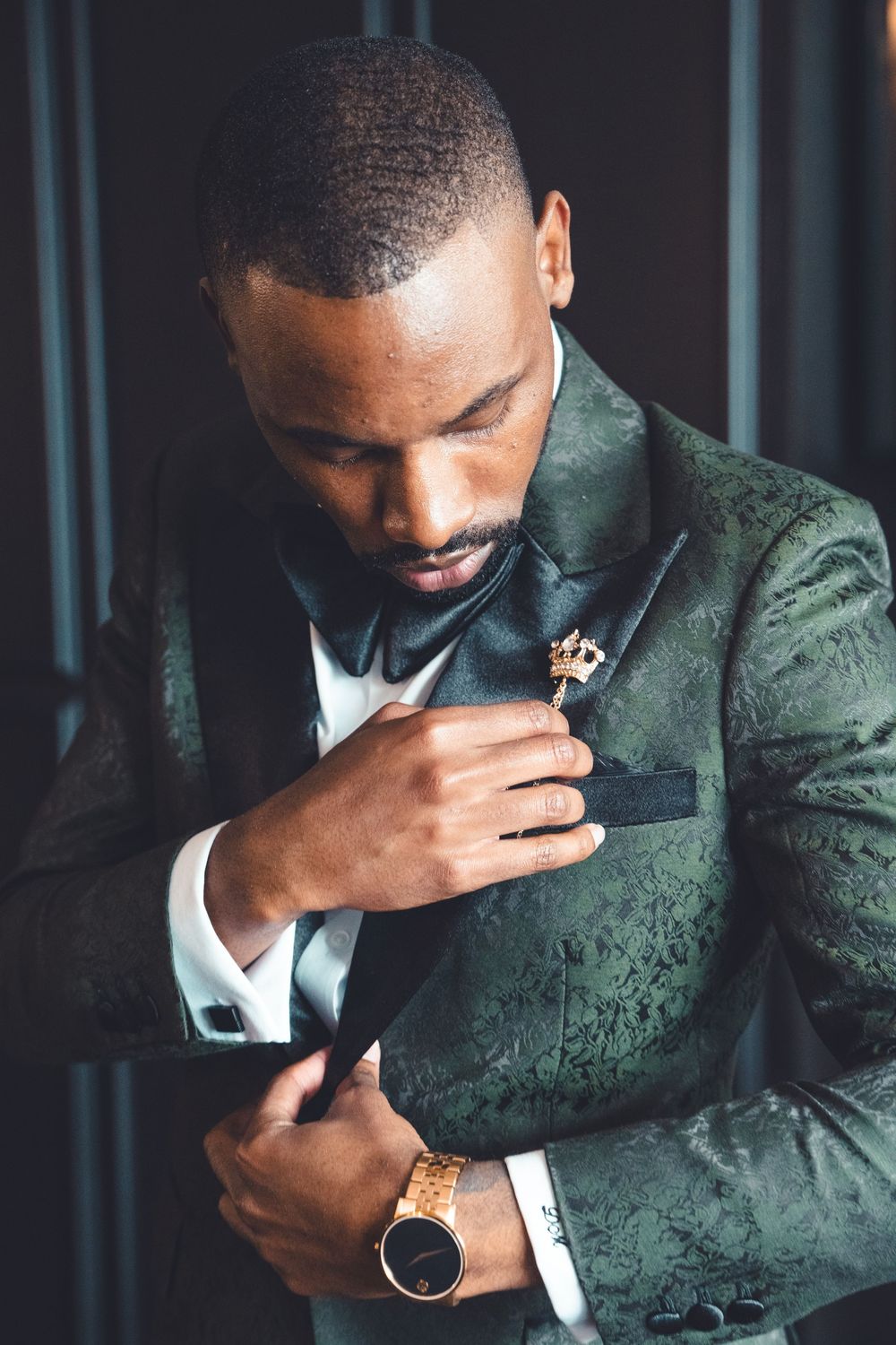 A person in an elegant emerald green suit adjusts their lapel flower while wearing a luxurious watch and white dress shirt.