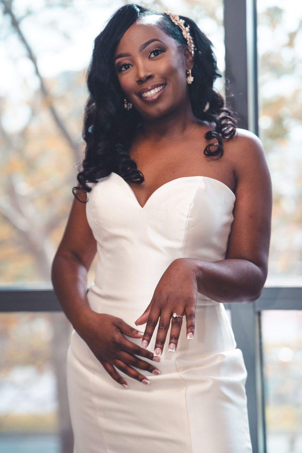 A bride in an elegant strapless white wedding gown showing off her ring in front of a window with autumn foliage.