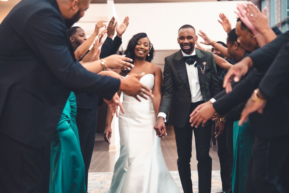 Joyful wedding couple walks through crowd of wedding party members in teal dresses clapping and celebrating.