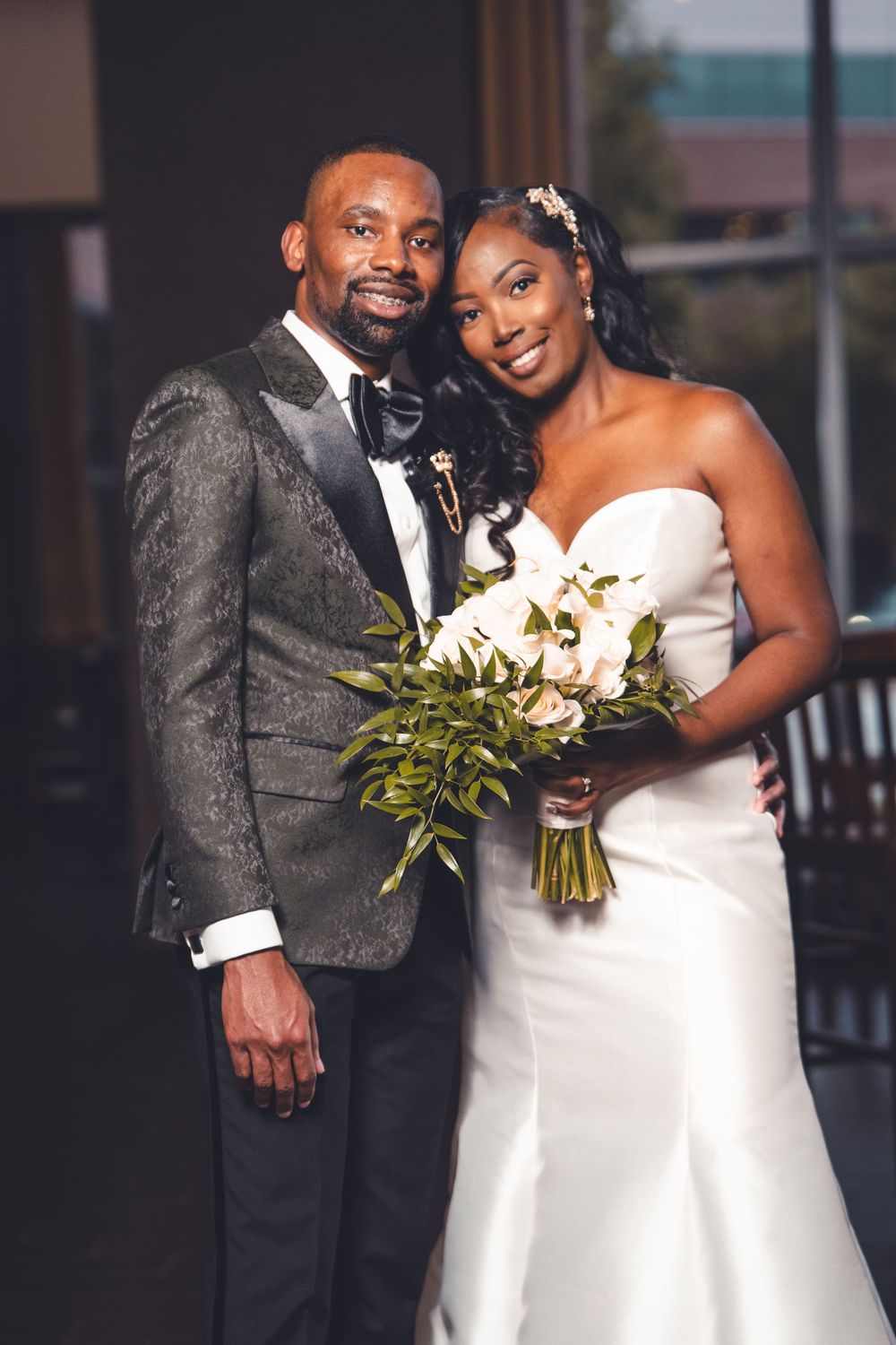 Wedding couple poses together in elegant attire with a beautiful white floral bouquet against an urban background.