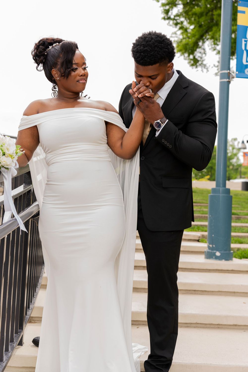 A wedding couple sharing a tender moment on outdoor stairs, with the bride in a white off-shoulder gown and groom in black suit.