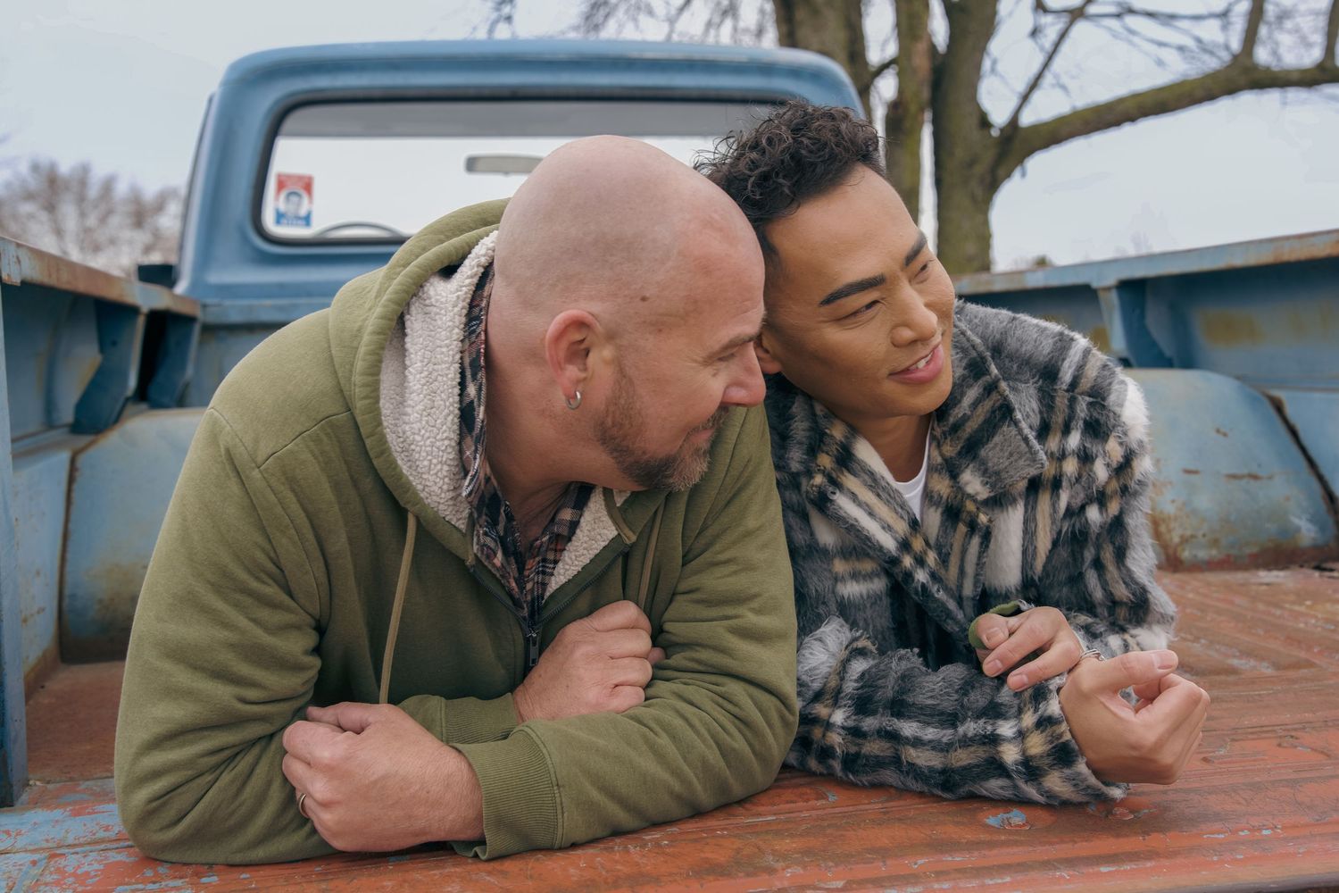 Two friends in casual attire leaning on a wooden surface with a vintage blue truck visible in the background.