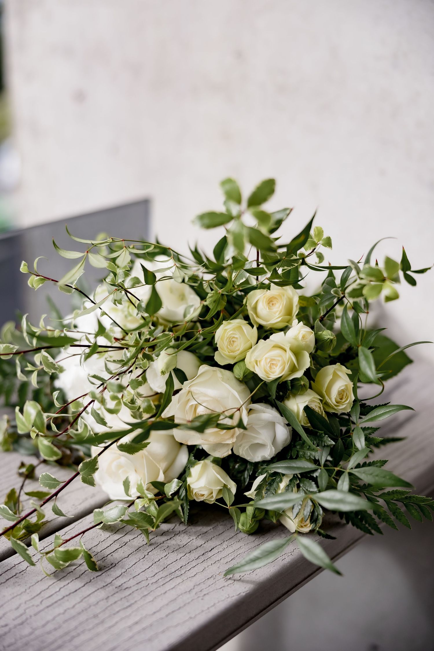 Elegant white rose wedding bouquet with delicate greenery and foliage resting on a gray wooden surface.