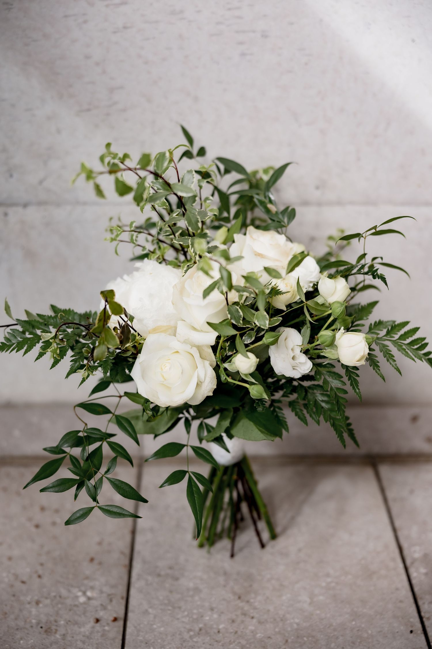Elegant white rose and greenery wedding bouquet with delicate foliage on concrete floor.