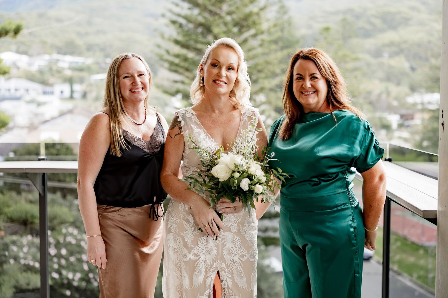 A bride in a white lace dress holds a white bouquet while posing with two guests on a balcony overlooking trees.