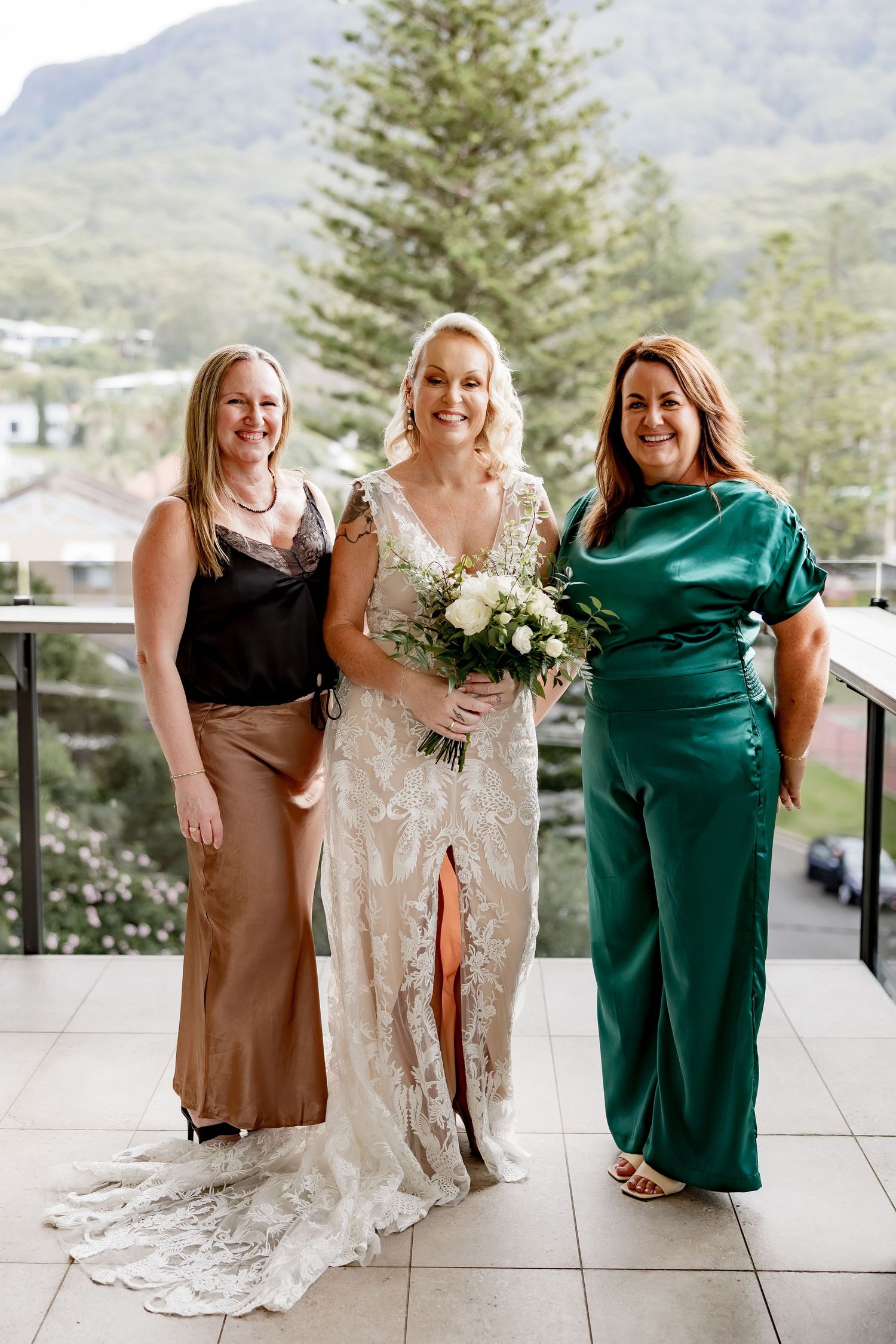Three friends pose on a balcony with scenic mountain views, wearing elegant formal attire for a wedding celebration.