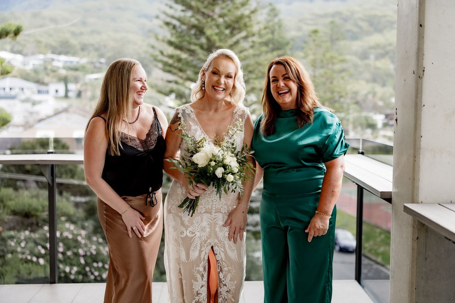 Three women in formal attire stand together on a balcony, with one in white holding a floral bouquet.