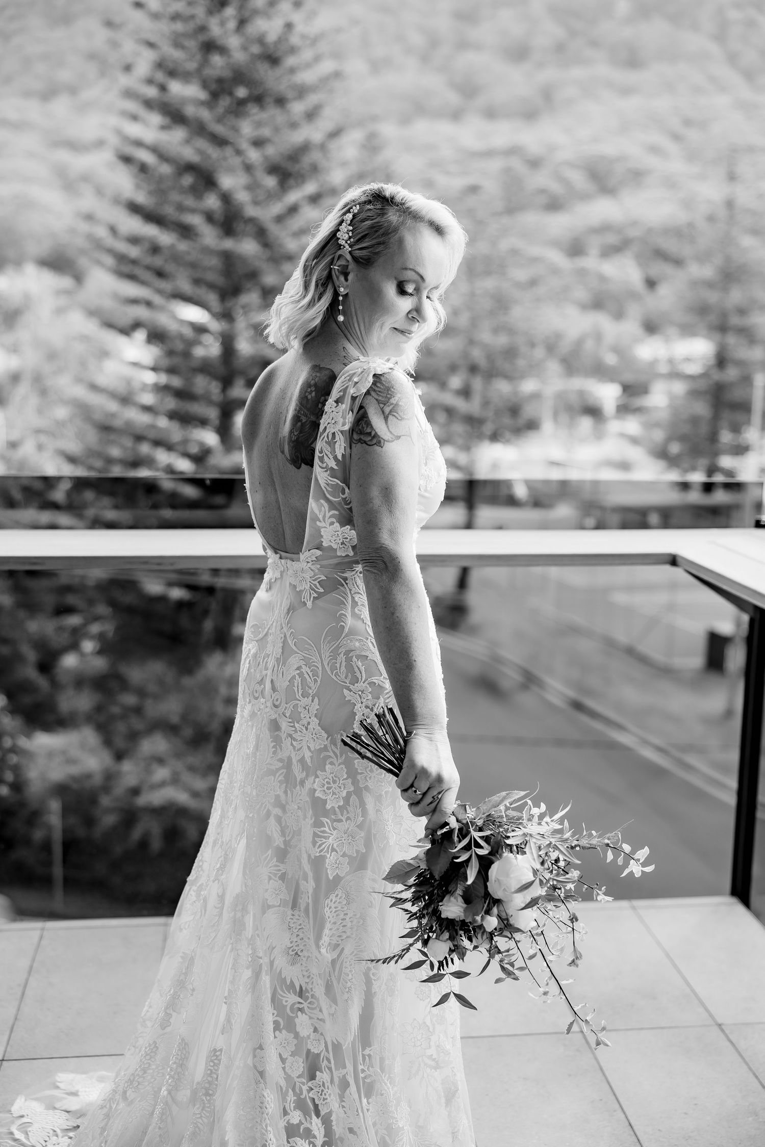 Bride in lace dress holding bouquet on outdoor balcony with mountain view in black and white.