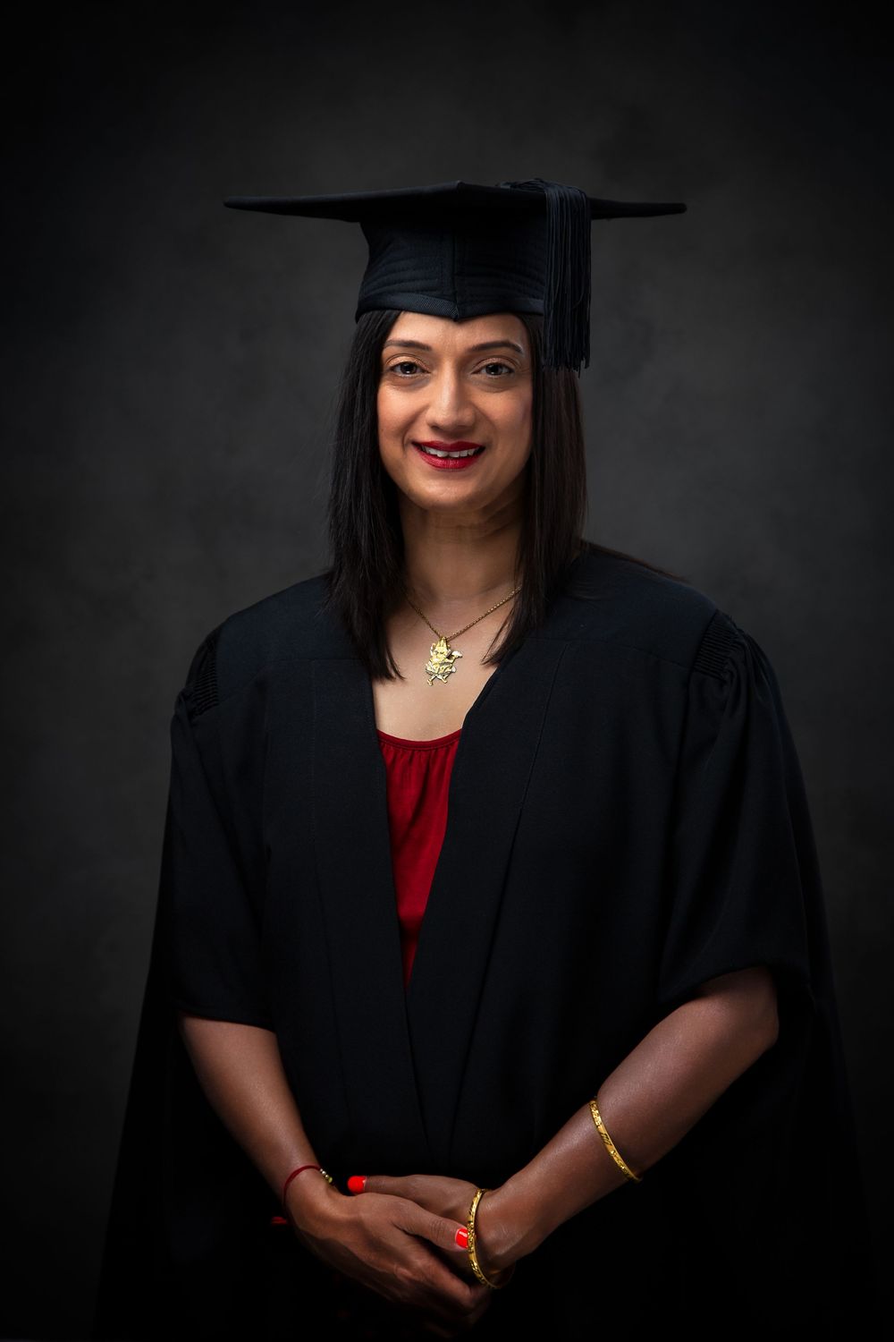 Graduate wearing academic regalia with cap and gown poses against dark background for formal portrait.