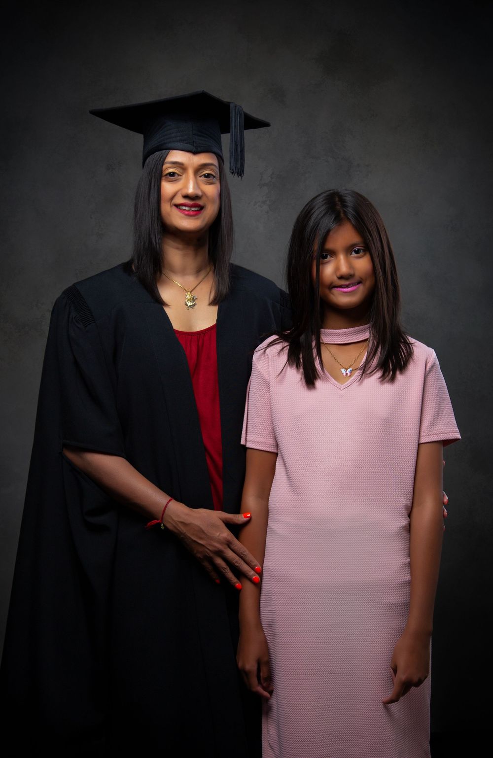 Graduate in black academic regalia poses with companion in pink dress against dark background.
