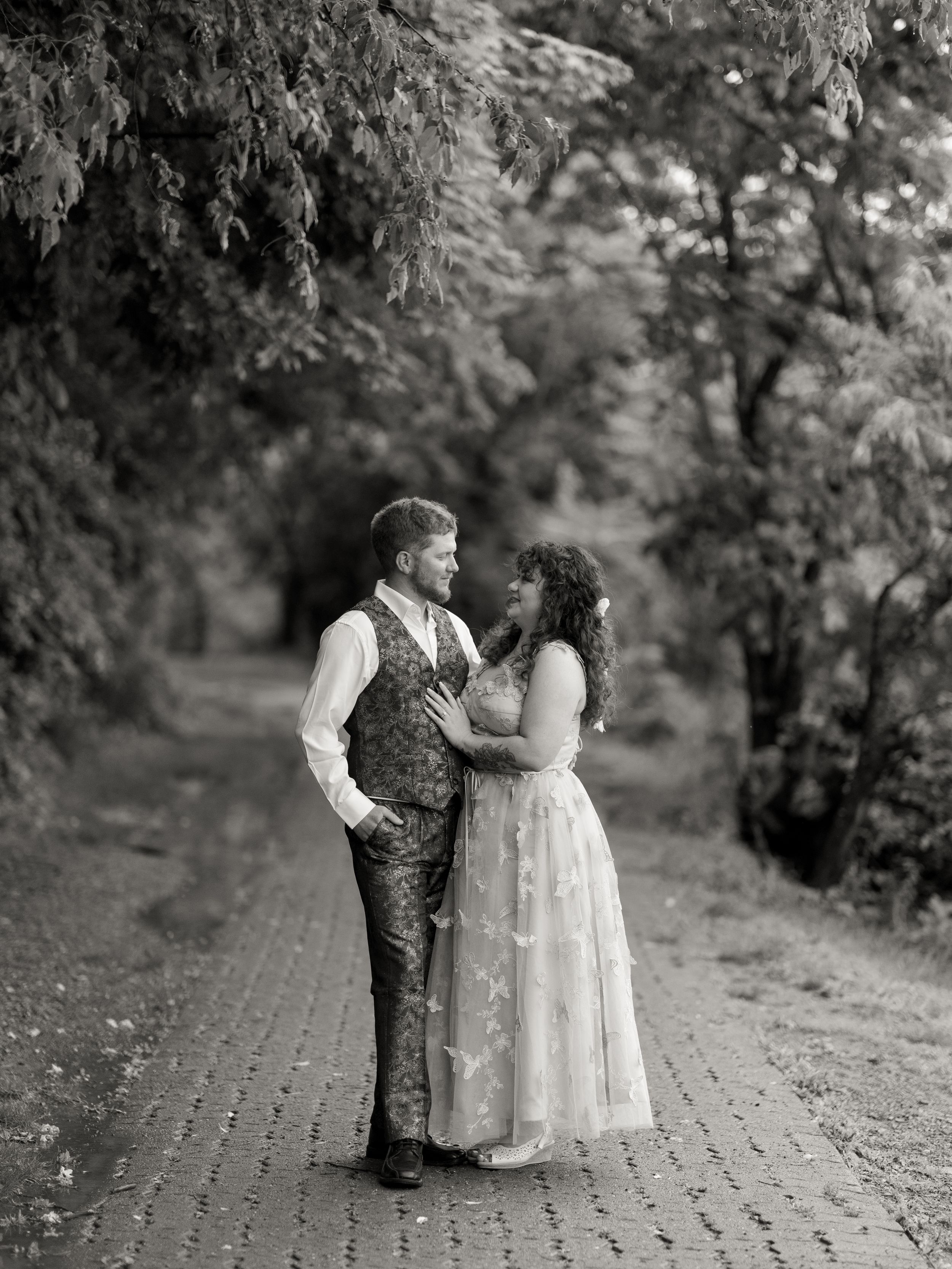 A romantic couple in vintage style attire sharing an intimate moment on a brick path surrounded by lush trees.