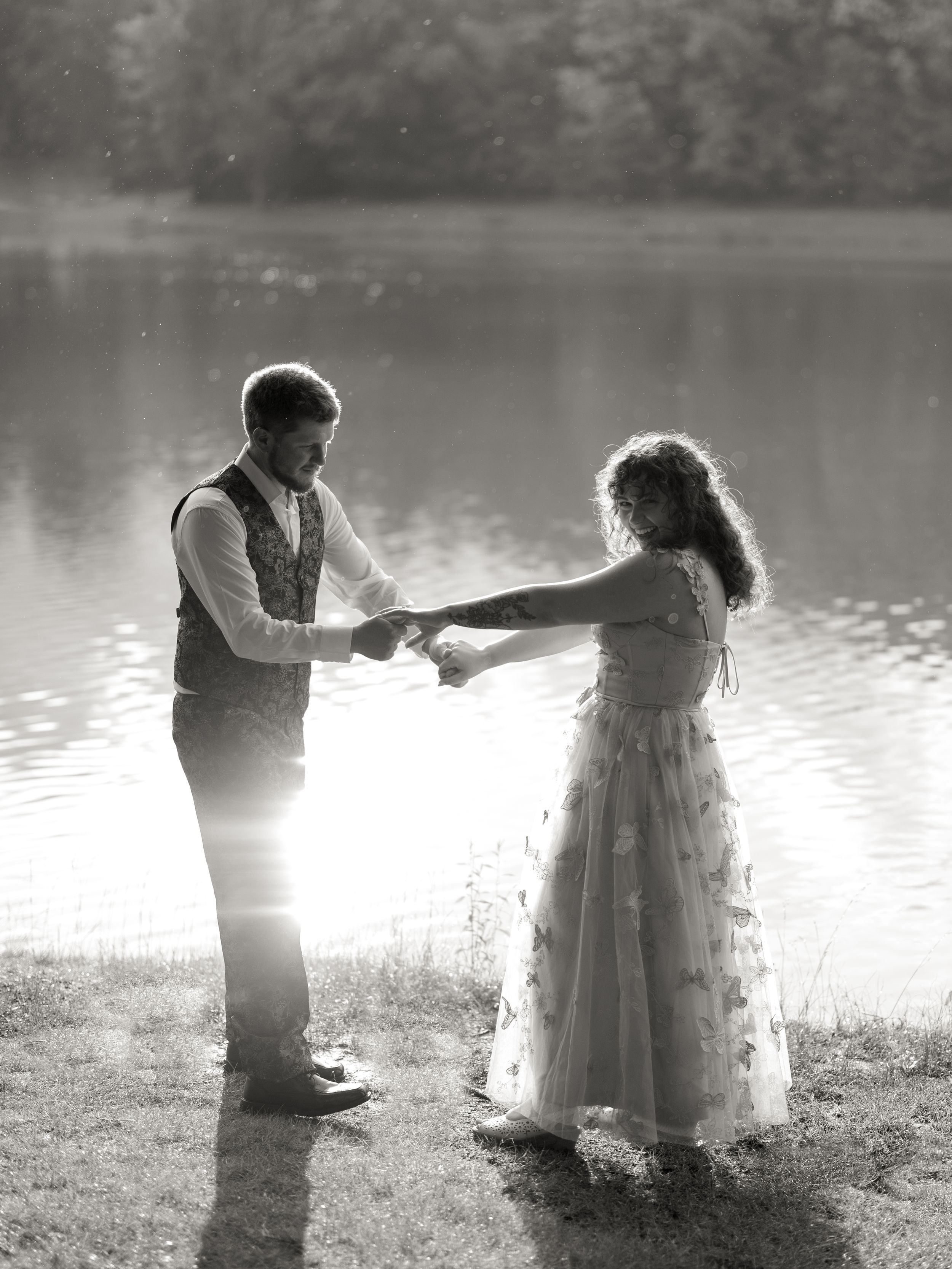 A romantic black and white photo of a couple dancing by a serene lake in formal vintage-inspired attire.