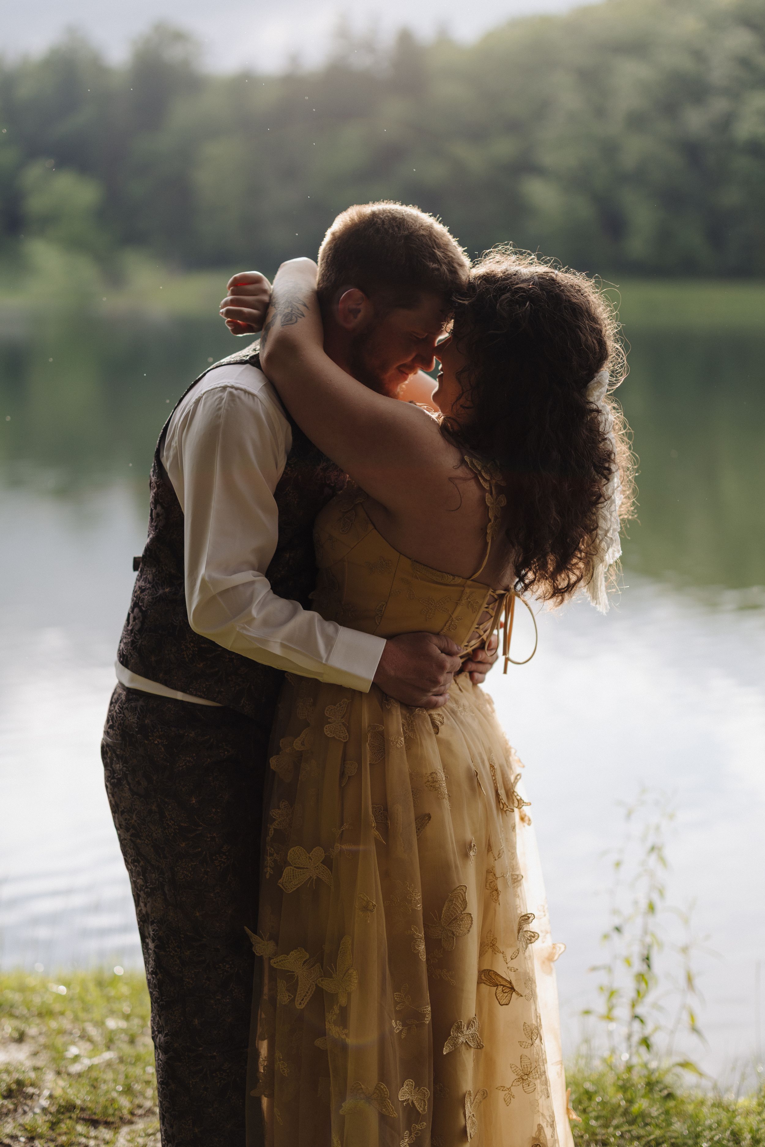 A couple embraces in formal attire by a tranquil lakeside with lush greenery in the background.