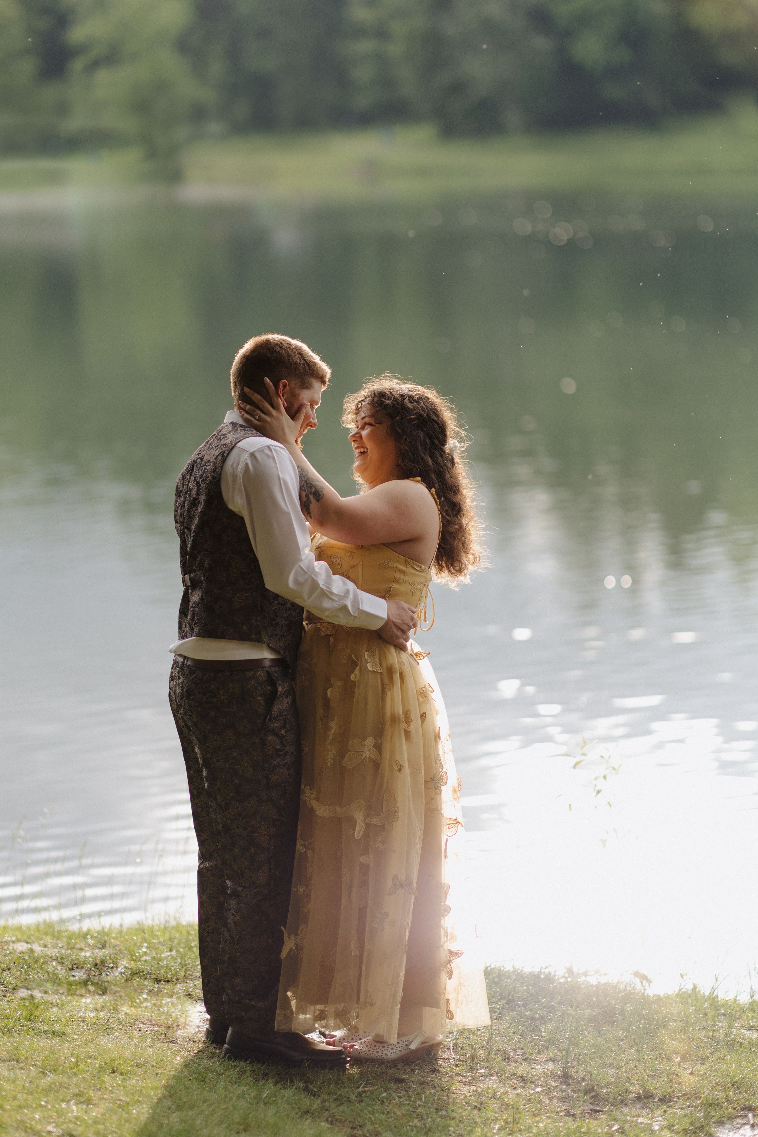 A couple in vintage-inspired attire embraces by a serene lake during a romantic outdoor photoshoot.