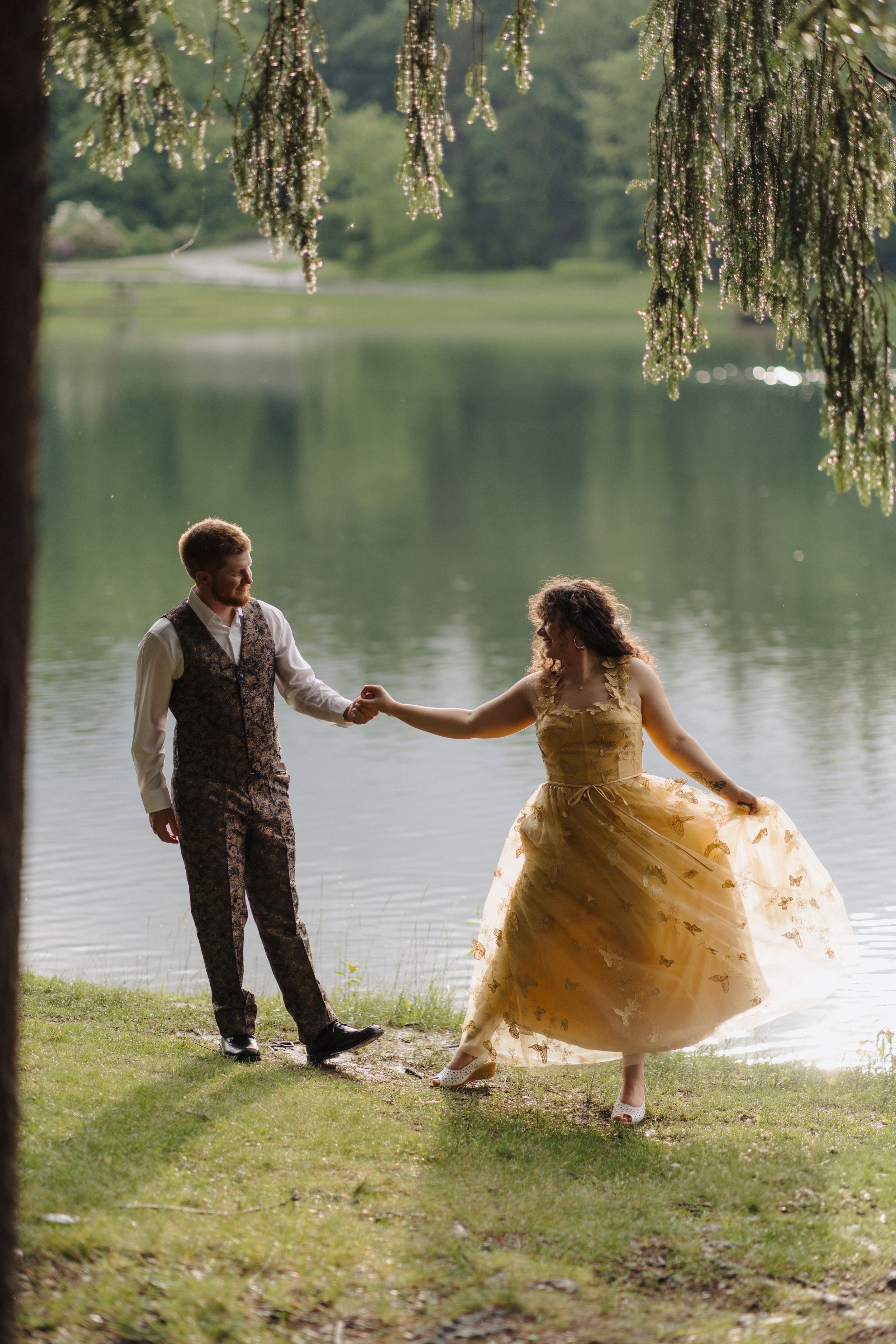 A couple in golden vintage clothing dance together on the grass beside a tranquil lake under willow branches.