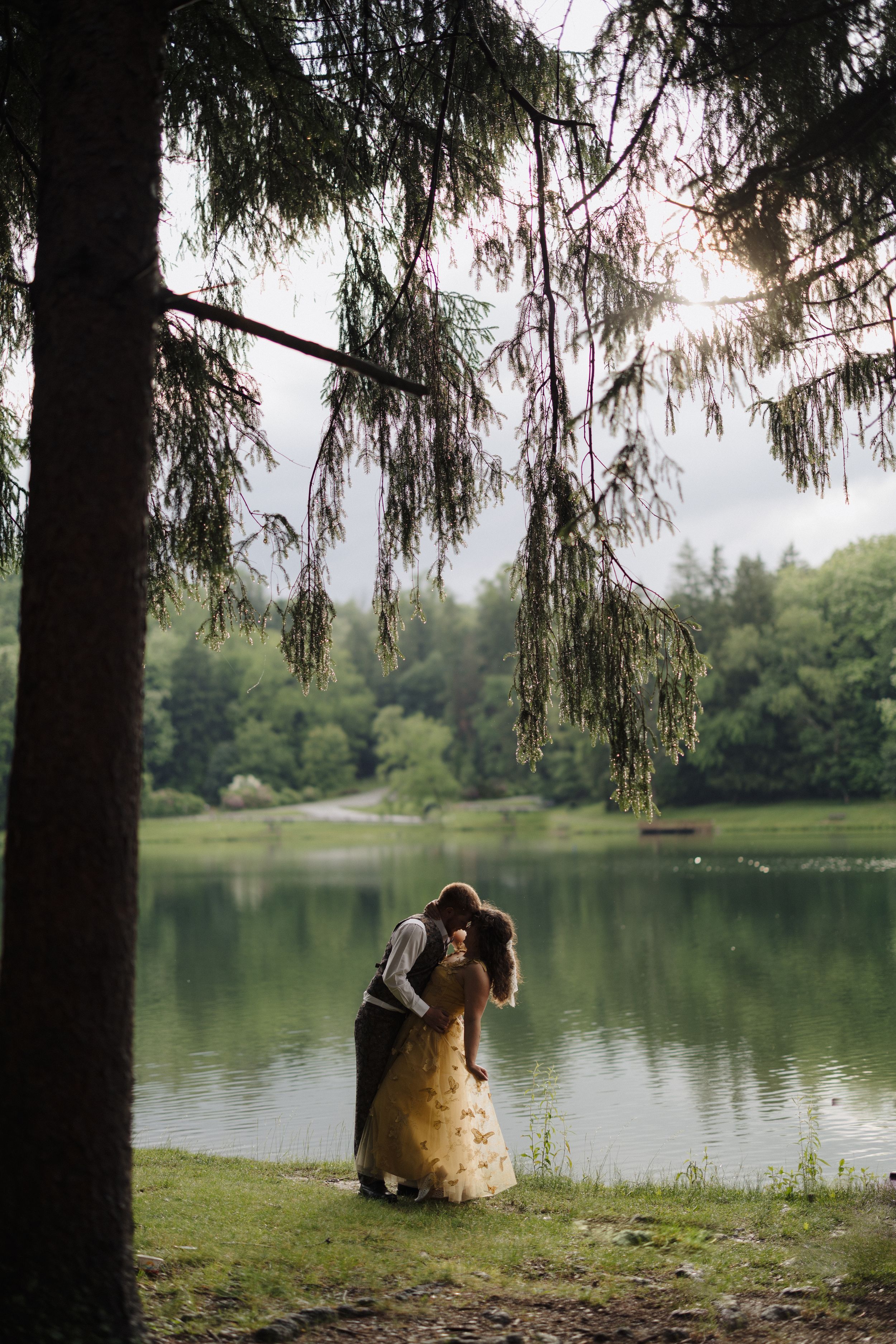 A couple embraces by a serene lake under drooping evergreen branches at sunset with reflective water in the background.