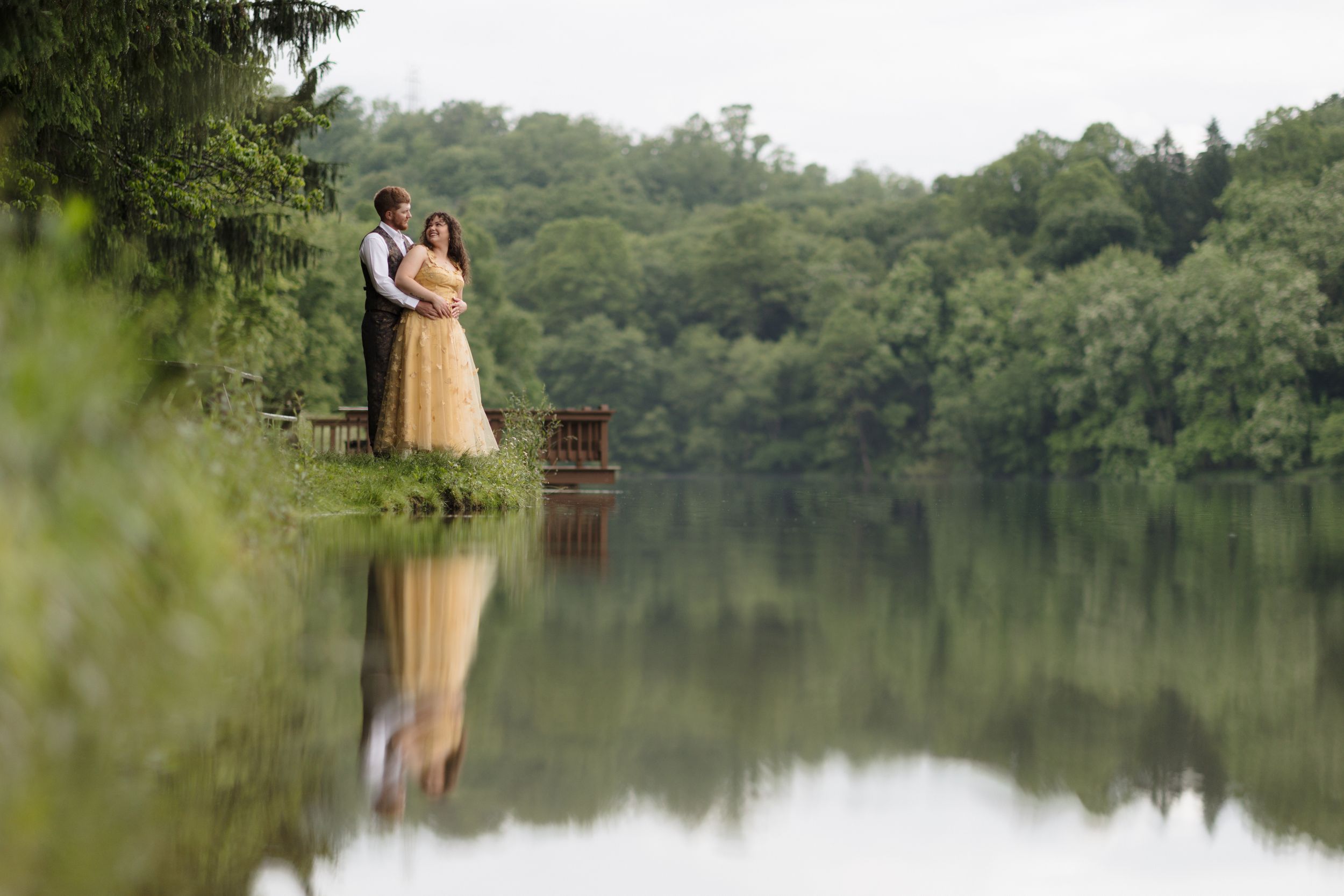 A couple in romantic poses stands by a calm lake with their reflection visible in the still water.