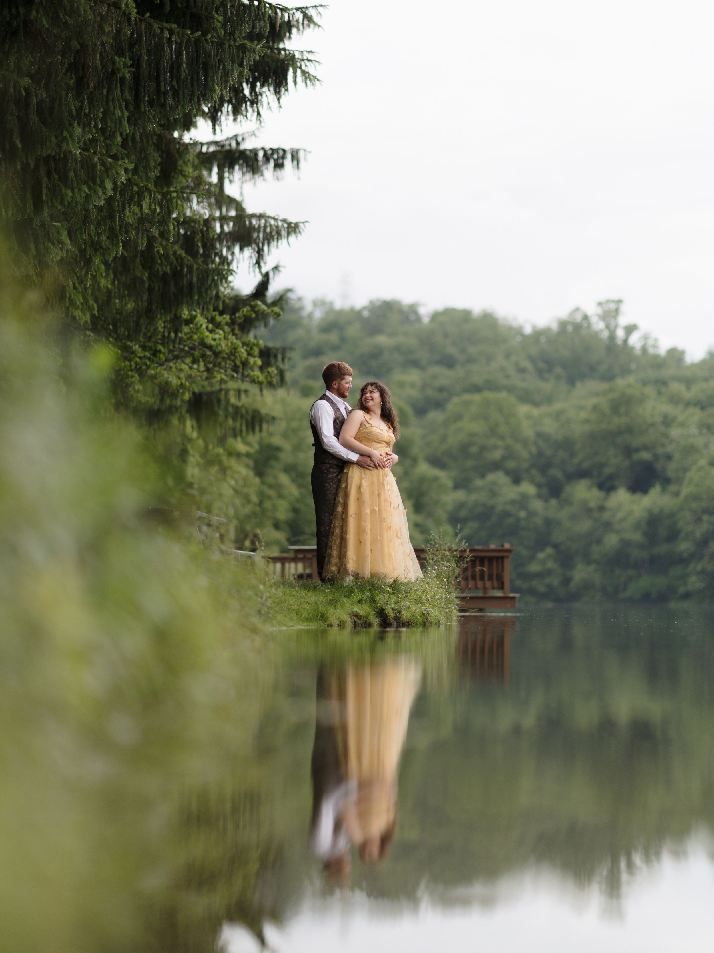 A couple in formal wear stands by a reflective lake with trees, their image mirrored in the calm water.