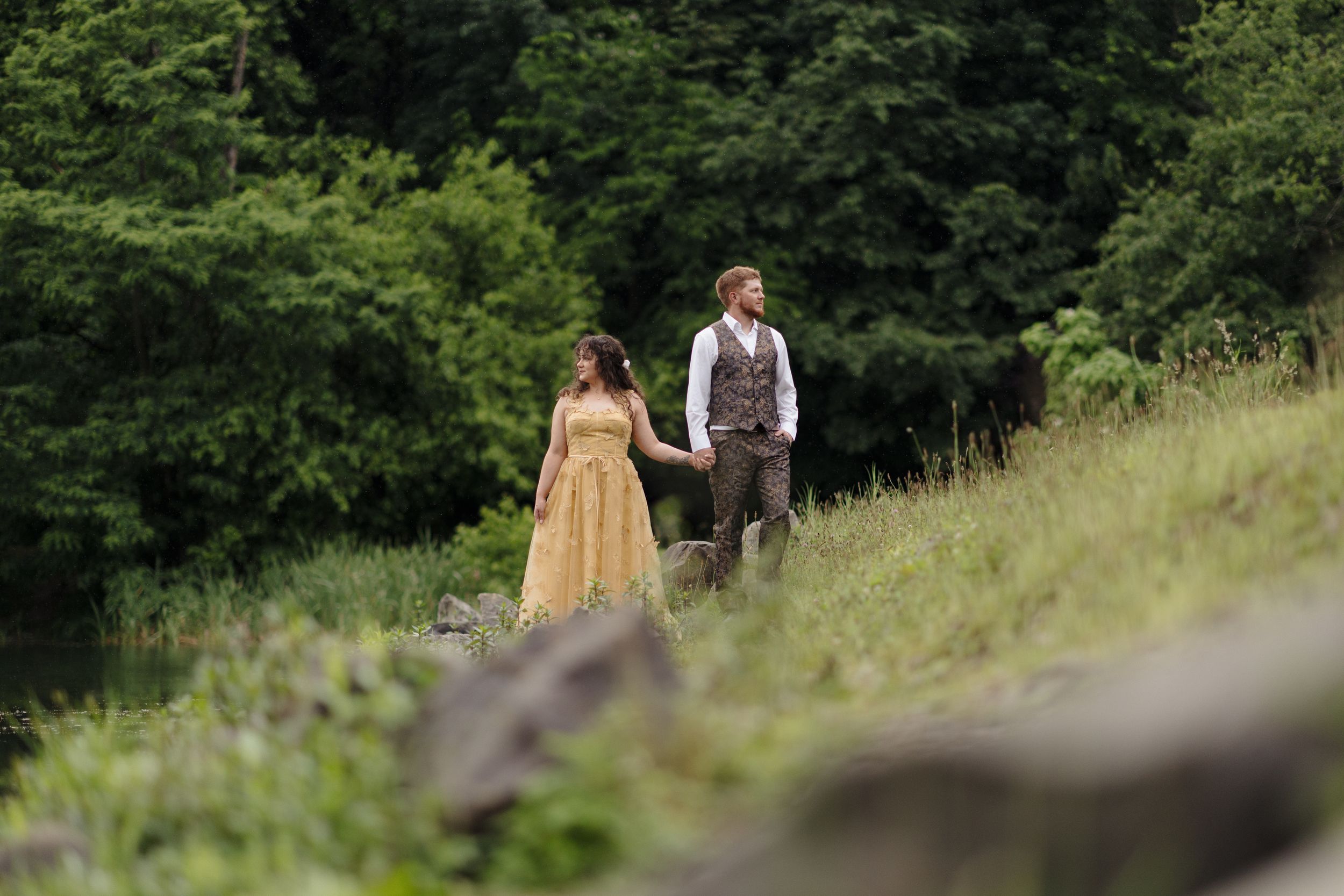 Two people in vintage clothing walking together on a grassy hillside surrounded by lush green forest.