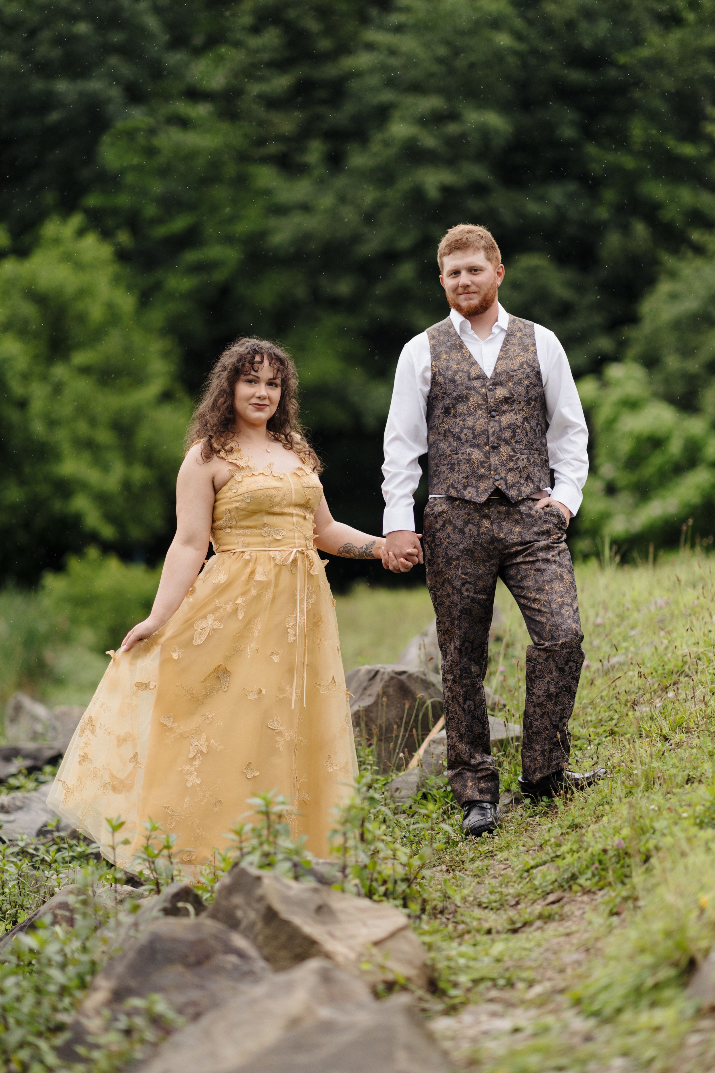 A couple stands on rocky terrain surrounded by greenery, with the woman in a gold dress and man in paisley vest.