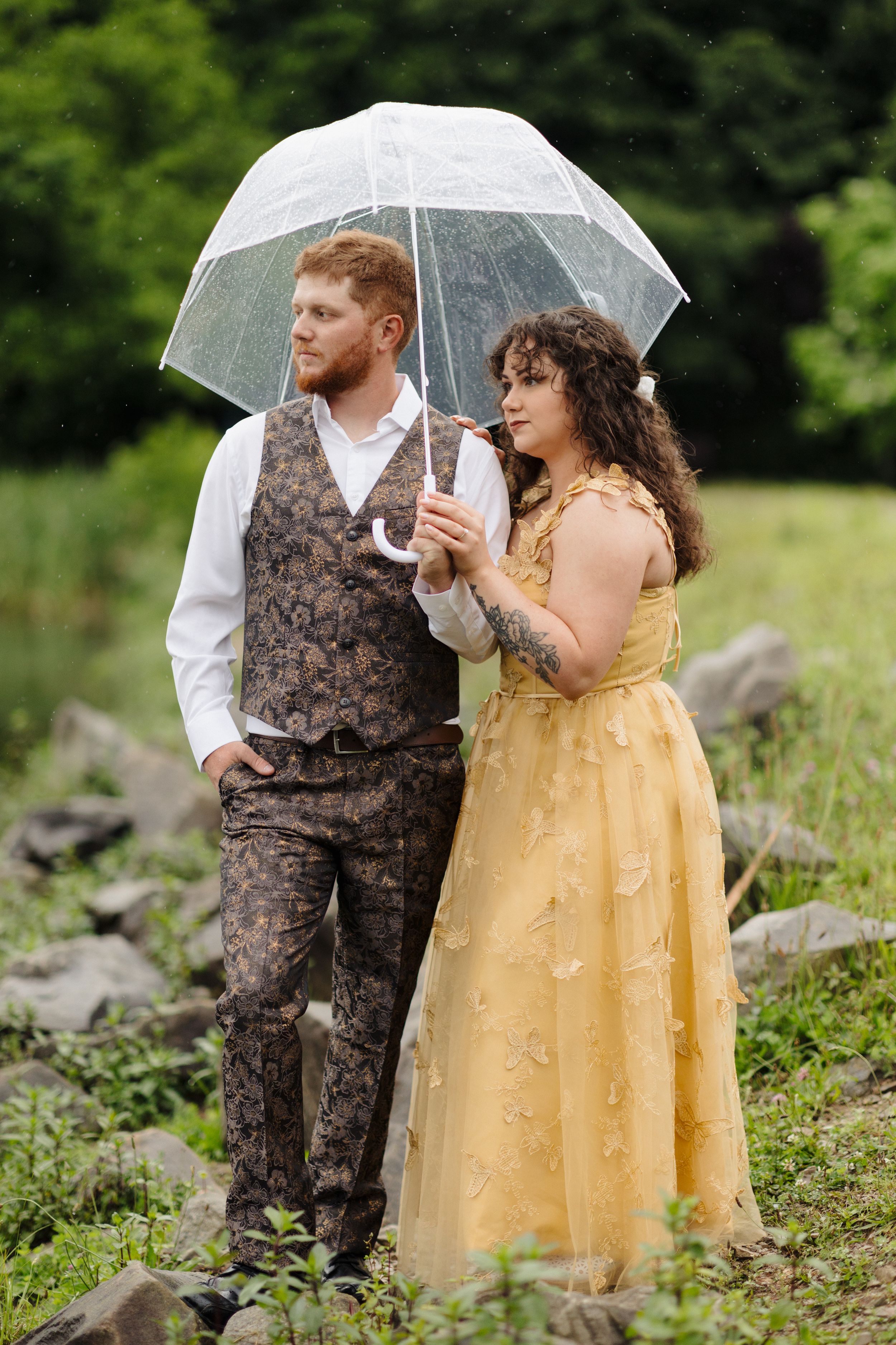 A couple stands together under a clear umbrella in a field, wearing vintage-inspired formal wear in brown and yellow.