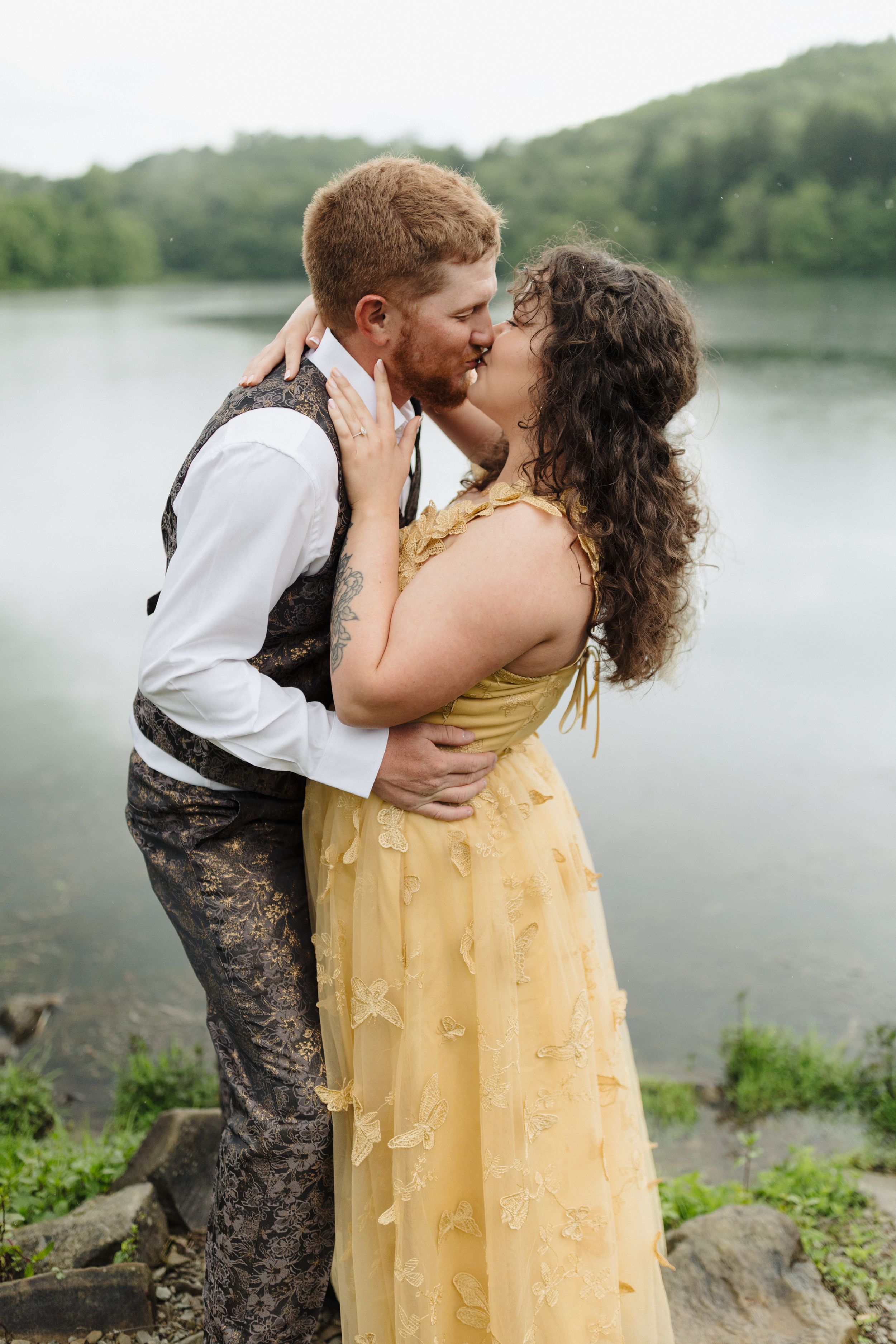 A couple in formal dress embracing by a serene lake, with the woman wearing a flowing yellow gown.