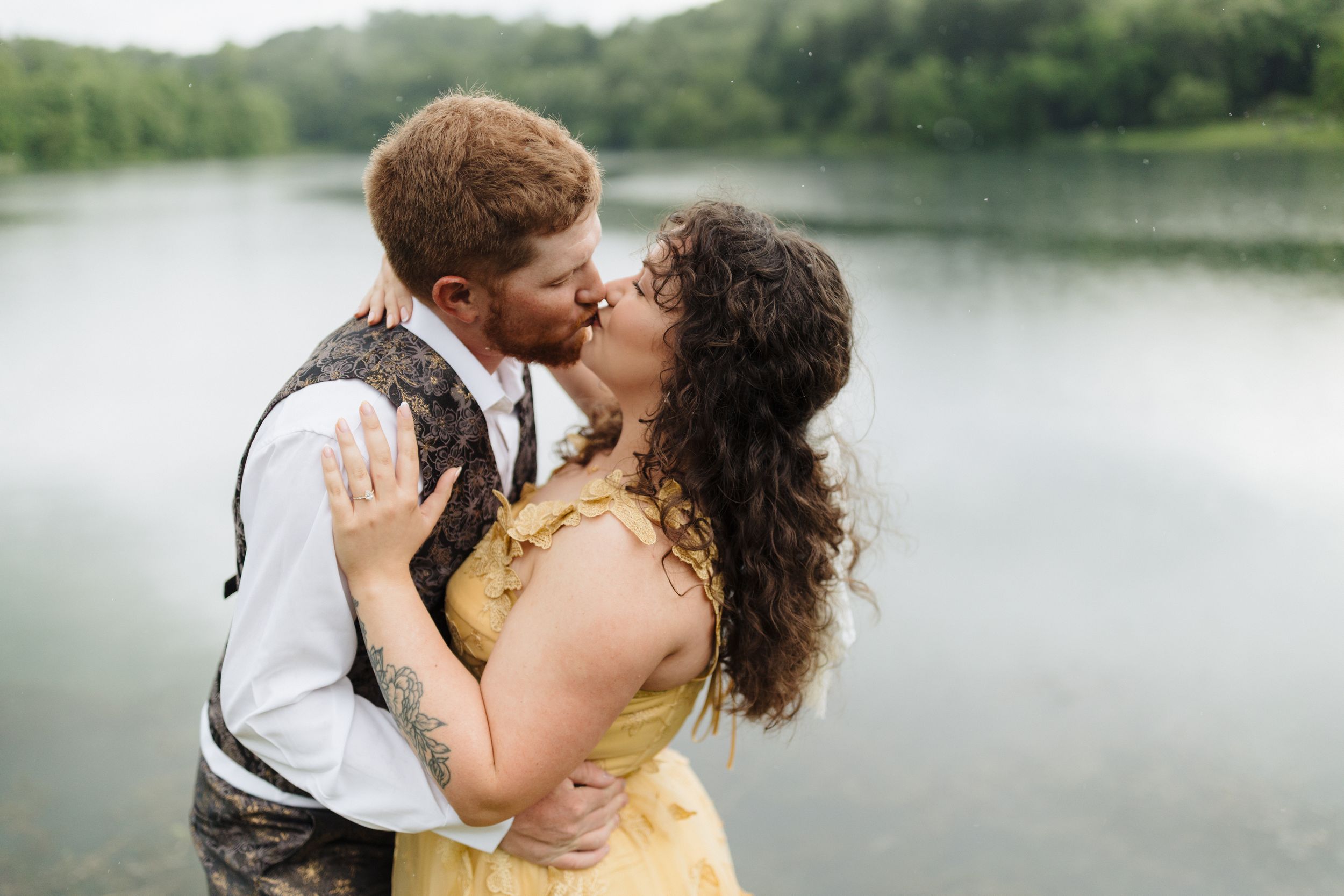 Two people in wedding attire embrace in a romantic kiss by a serene lake with lush green trees in the background.