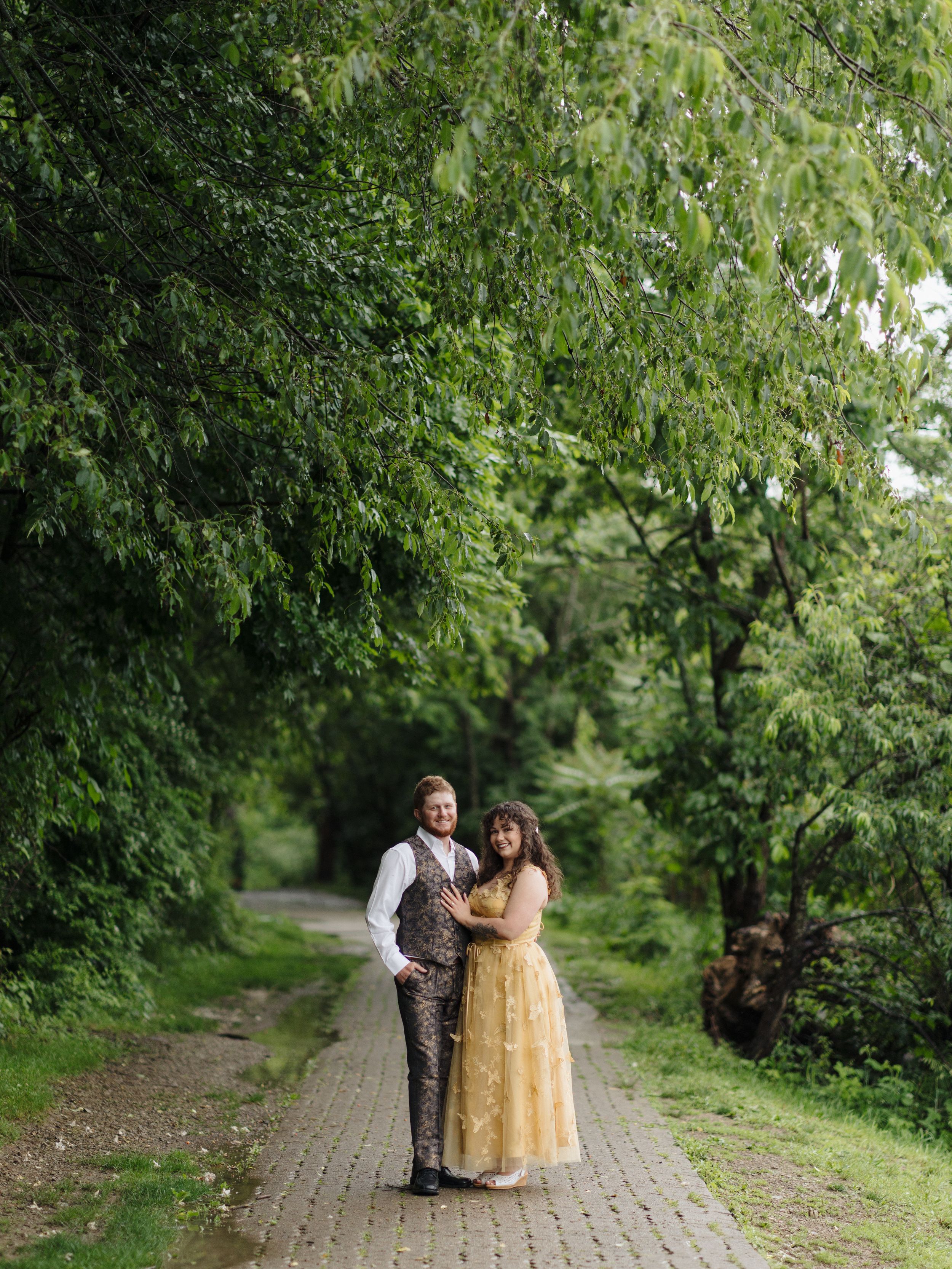 A couple in formal attire poses on a tree-lined path, she in a yellow gown and he in a brown vest and slacks.