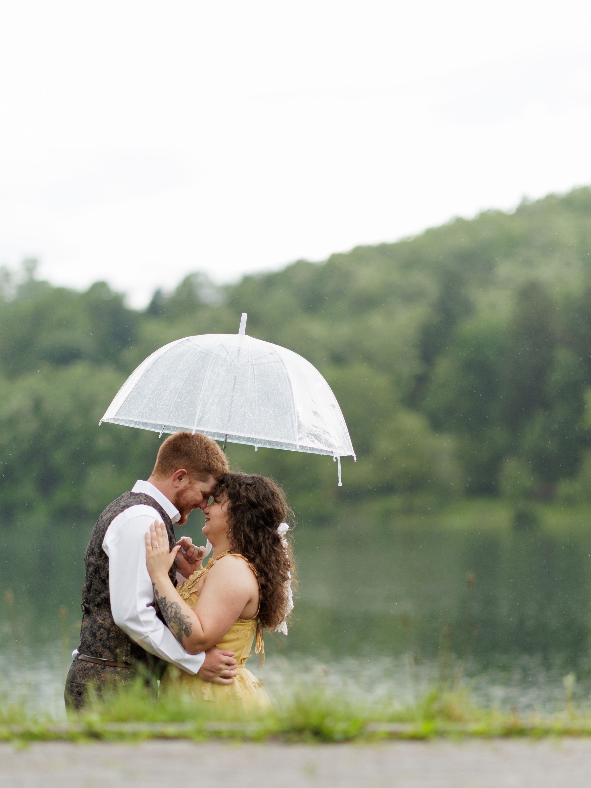 A romantic couple shares an intimate moment under a white umbrella by a serene lake surrounded by trees.